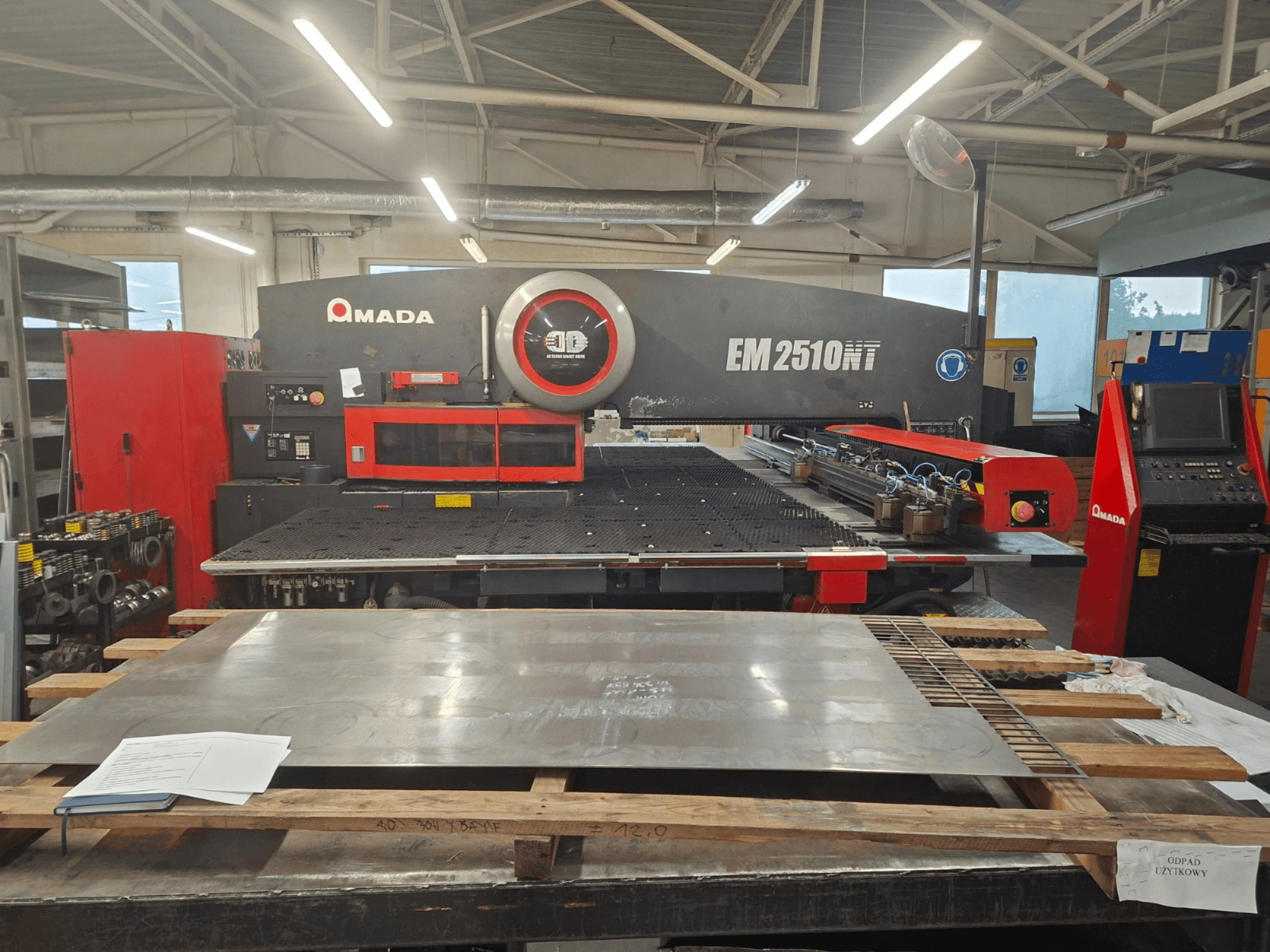 AMADA EM 2510NT perforatora preses priekšējais skats ar darbnīcā redzamu metāla loksni un vadības paneli.