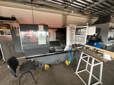 Haas CNC virpa (ST-10 modelis) darbnīcā, skats no sāniem, kurā redzamas vadības ierīces, instrumenti uz darbgalda un krēsls blakus.