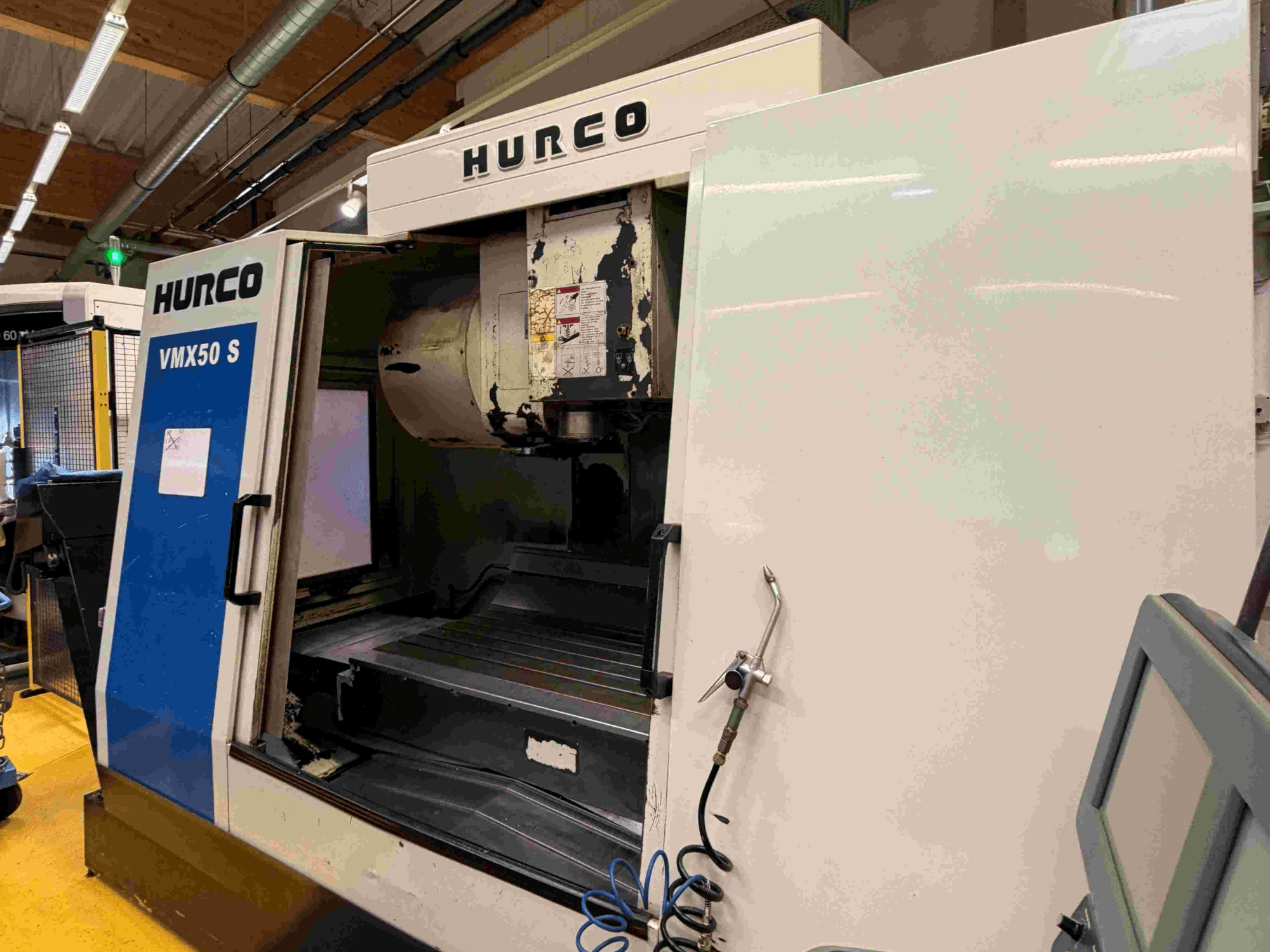 Hurco VMX50 S vertikālais apstrādes centrs, skats no sāniem, redzamas atvērtas durvis un vadības panelis.