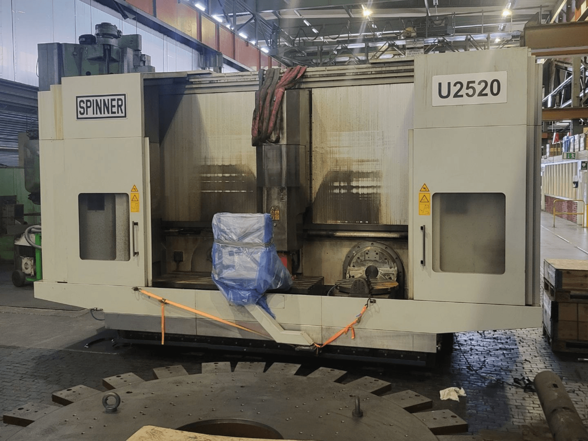Vertikālais apstrādes centrs SPINNER U5-2520, redzams no priekšpuses, ar pārklātu galdu un drošības uzlīmēm.