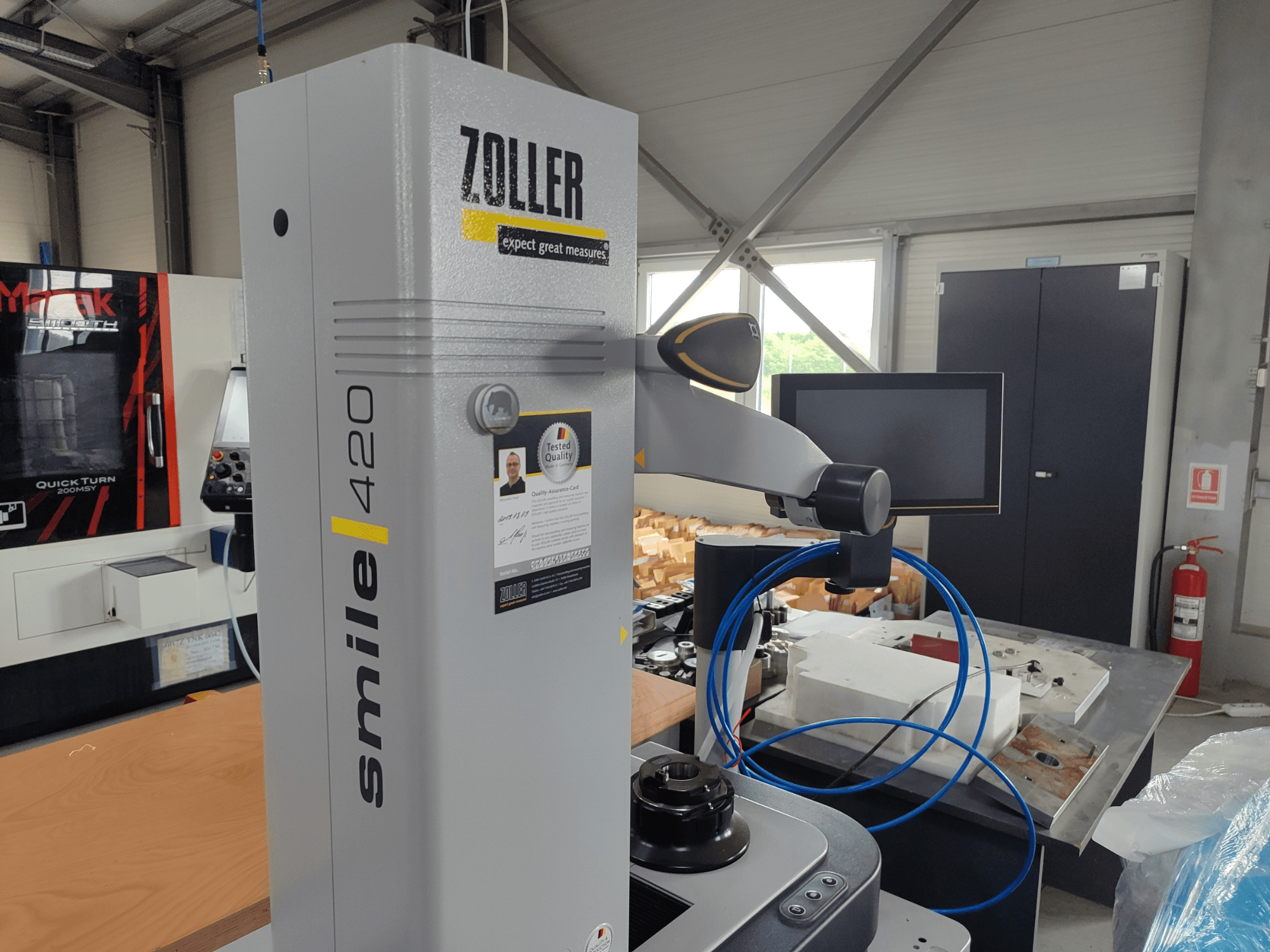 ZOLLER smile 420 mērierīce ar monitoru, skats no sāniem darbnīcā, ar zilām šļūtenēm un iekārtām fonā.
