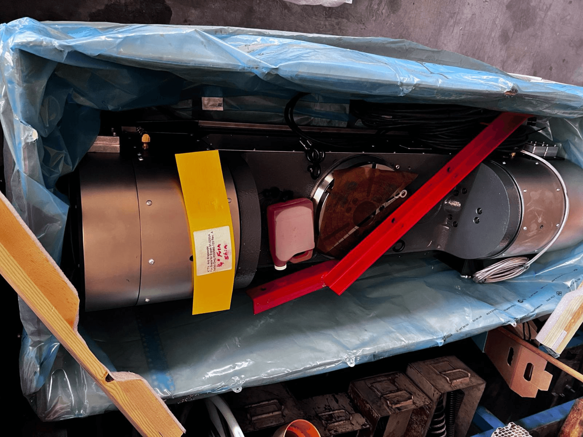 Motora ierīce daļēji redzama iepakojumā, tai ir sudraba ārpuse, krāsaini mērinstrumenti un sarkana atbalsta skava.