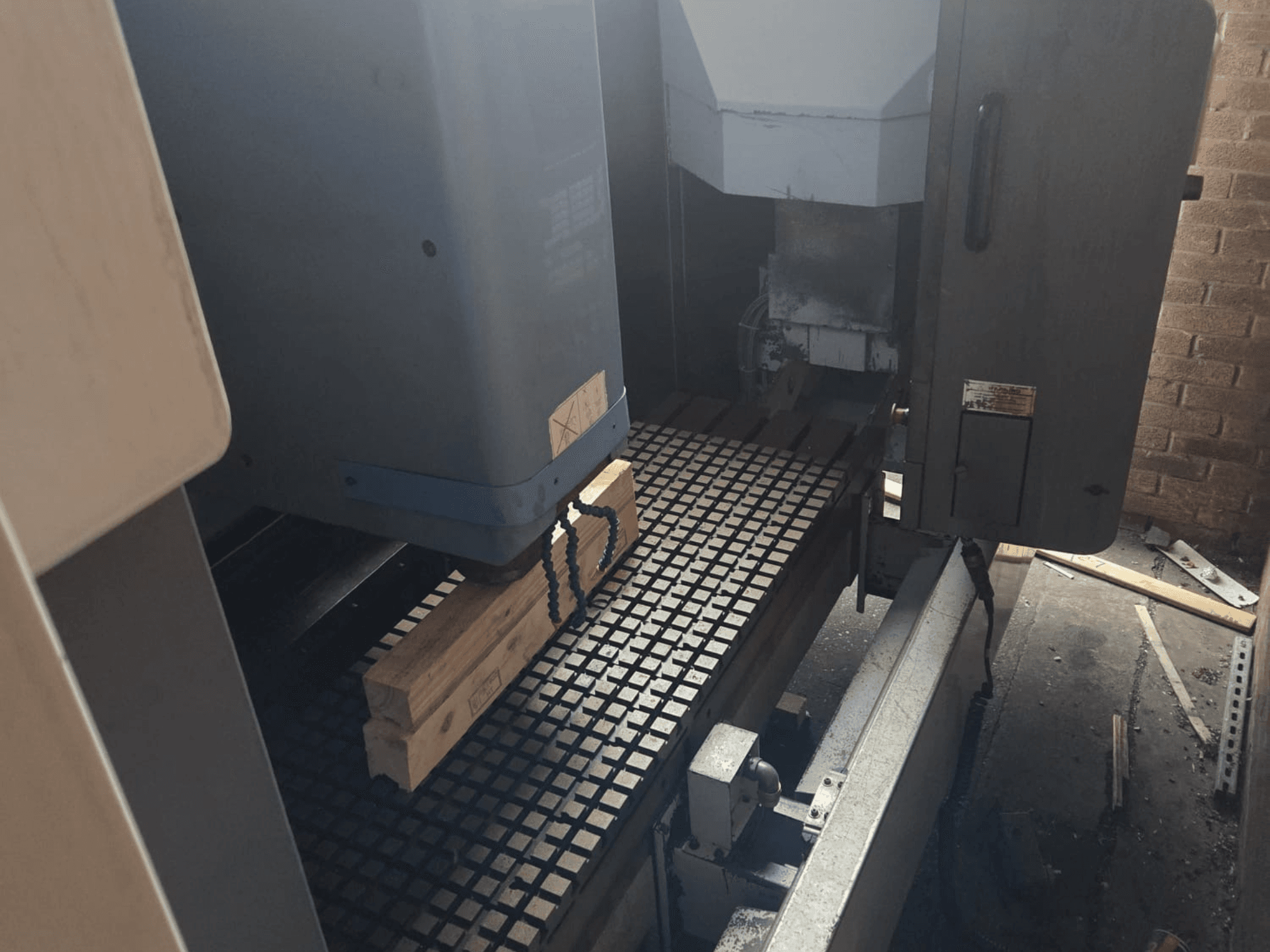 CNC mašīnas priekšējais skats ar koka dēļiem uz režģa galda, ar sānos redzamajiem instrumentiem un vadības ierīcēm.