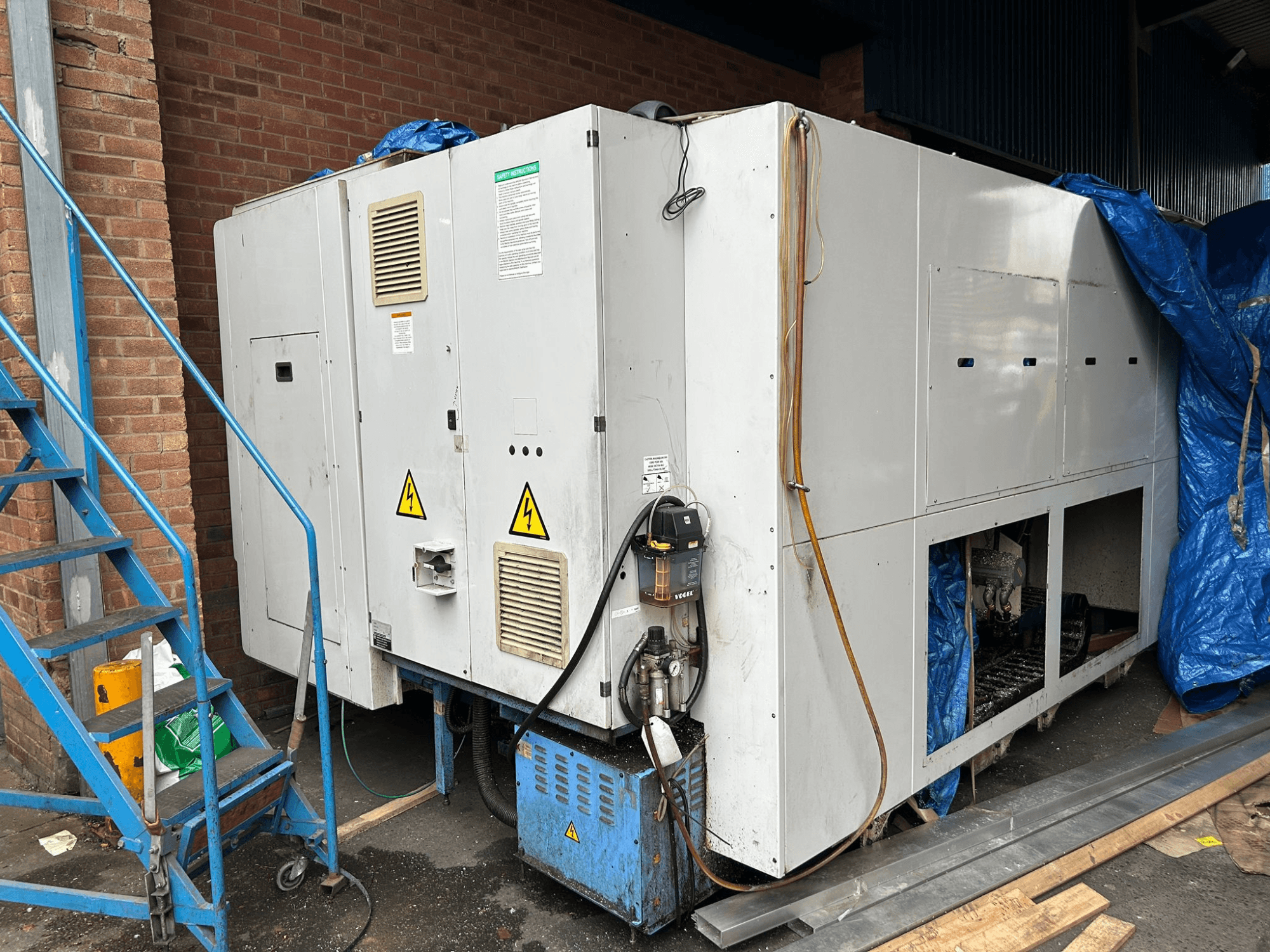 Skats no sāniem uz lielu rūpniecisku dzesēšanas iekārtu, zīmols un modelis neskaidrs, redzami elektriskie savienojumi, brīdinājuma zīmes un zils brezenta pārsegs.