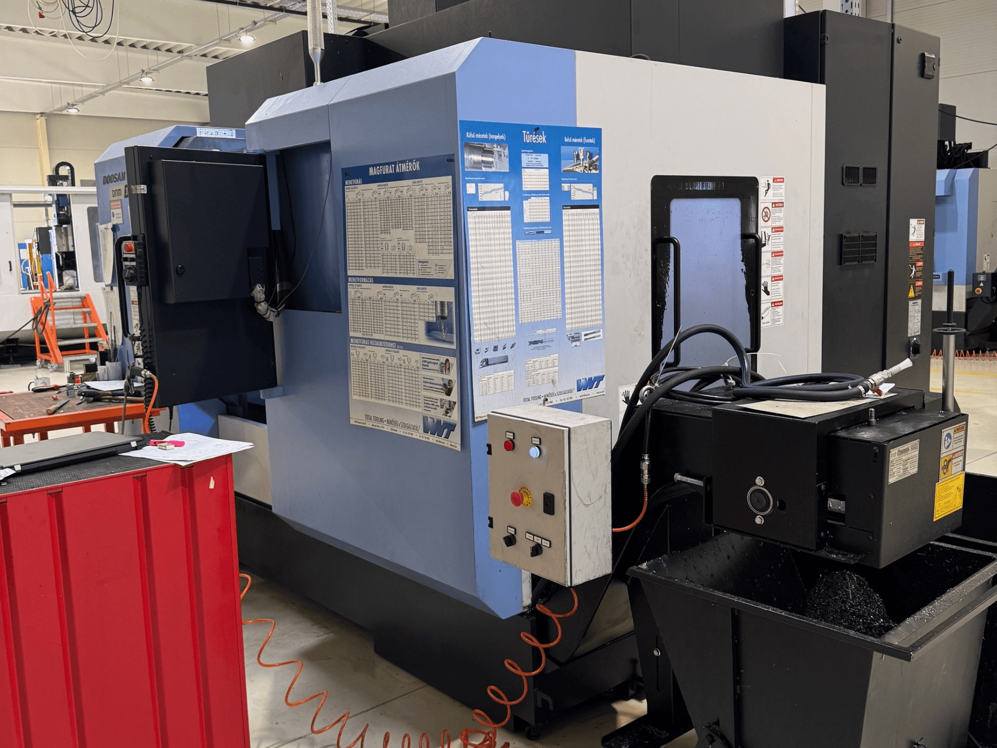 DOOSAN DNM350/5AX frēzēšanas un virpošanas centrs, sānskats, kurā redzams vadības panelis un dzesēšanas šķidruma tvertne, kā arī darba detaļas.