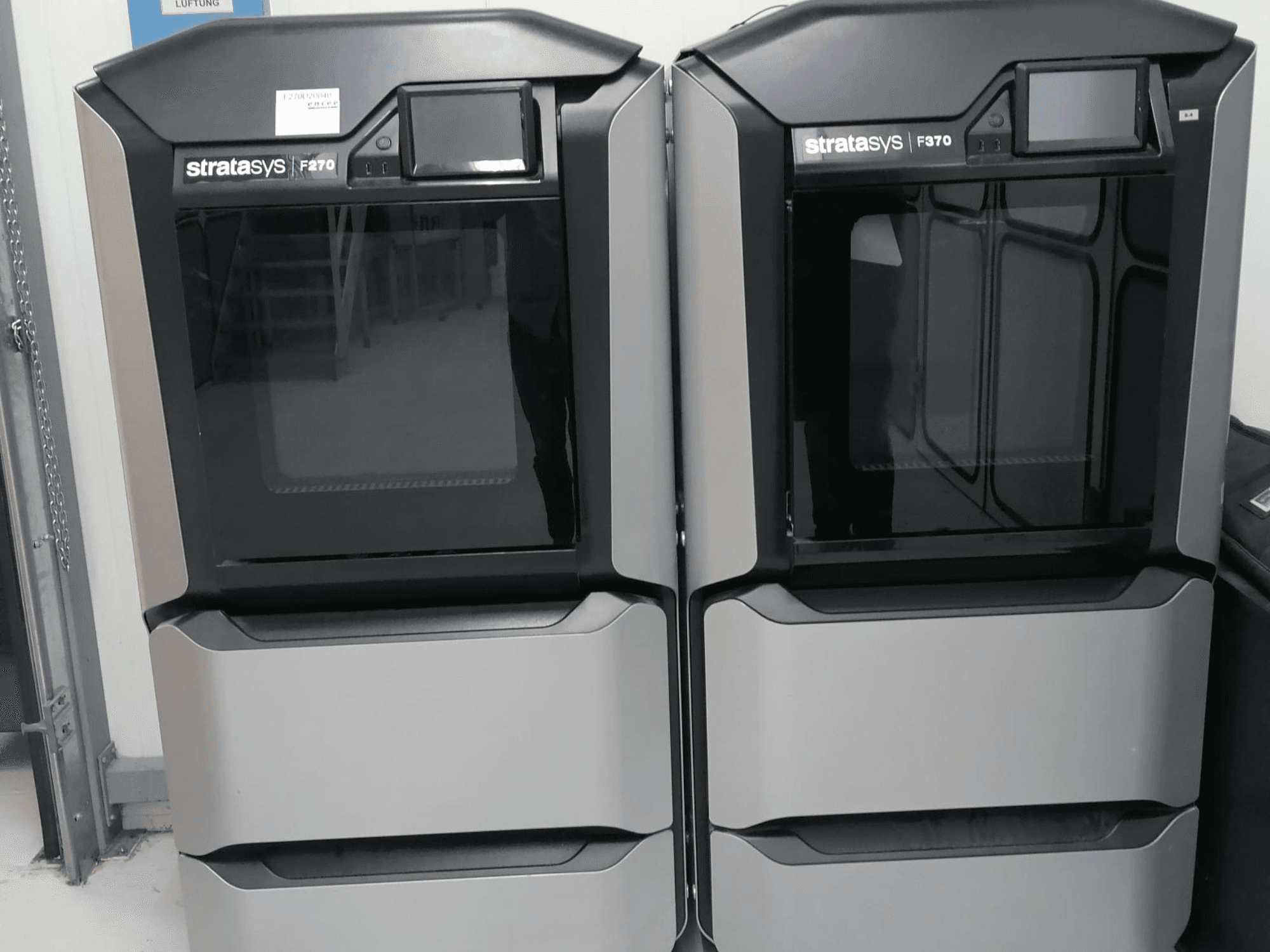 Stratasys F270 plastmasas 3D printera priekšējais skats, kurā redzamas divas iekārtas ar gludu pelēku ārpusi un digitālo displeju.
