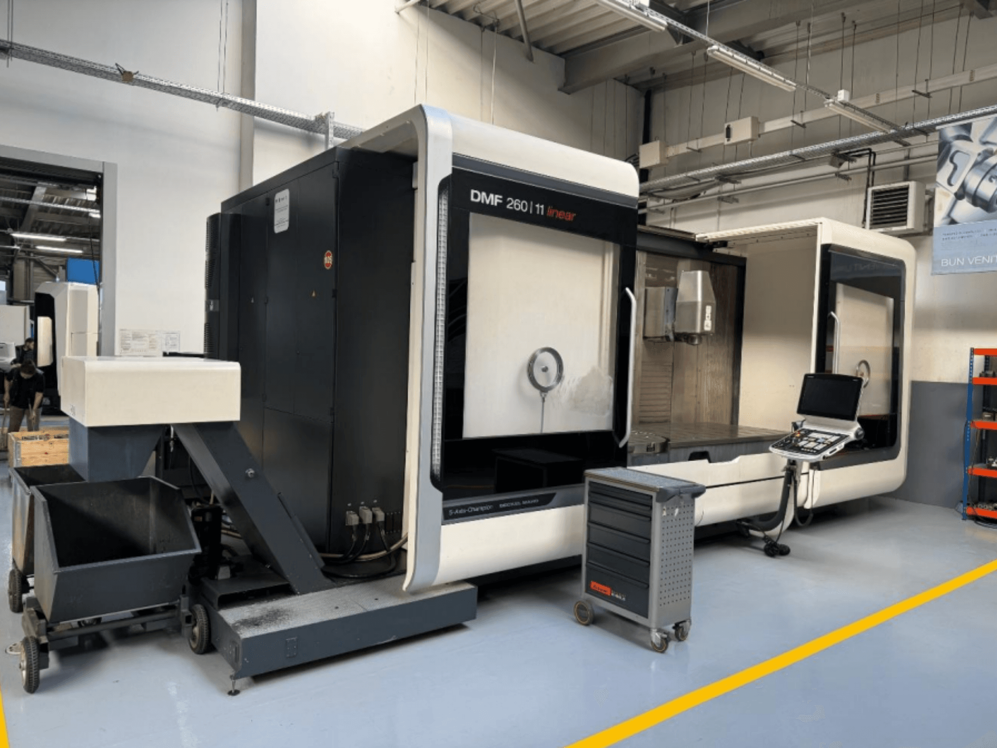 DMG MORI DMF 260 Lineārā frēzēšanas iekārta no priekšpuses ar instrumentu vadības paneli un pārvietojamo instrumentu ratiņu blakus.