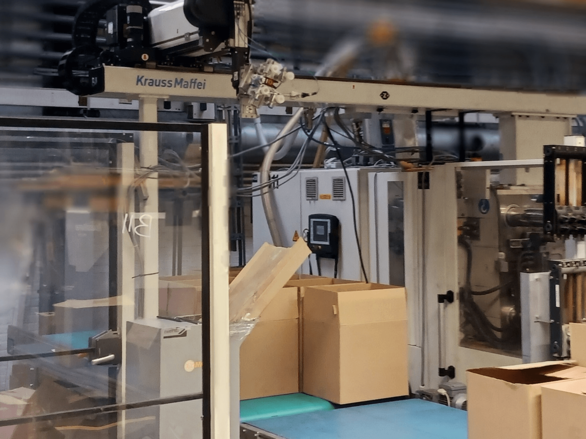 Krauss Maffei automatizācijas sistēma rūpnīcā, skats no priekšpuses, kurā redzamas kastes uz konveijera un darbojošās iekārtu sastāvdaļas.