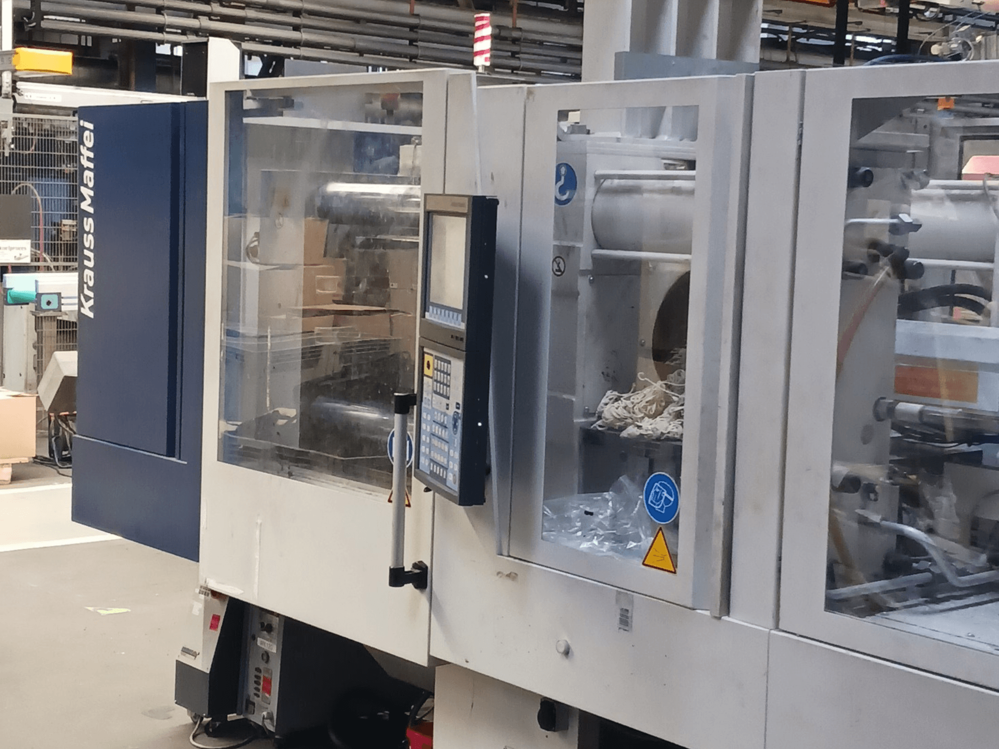 KraussMaffei injekcijas formēšanas iekārta, skats no sāniem, fonā redzams vadības panelis, iekārtas un iepakojuma materiāli.