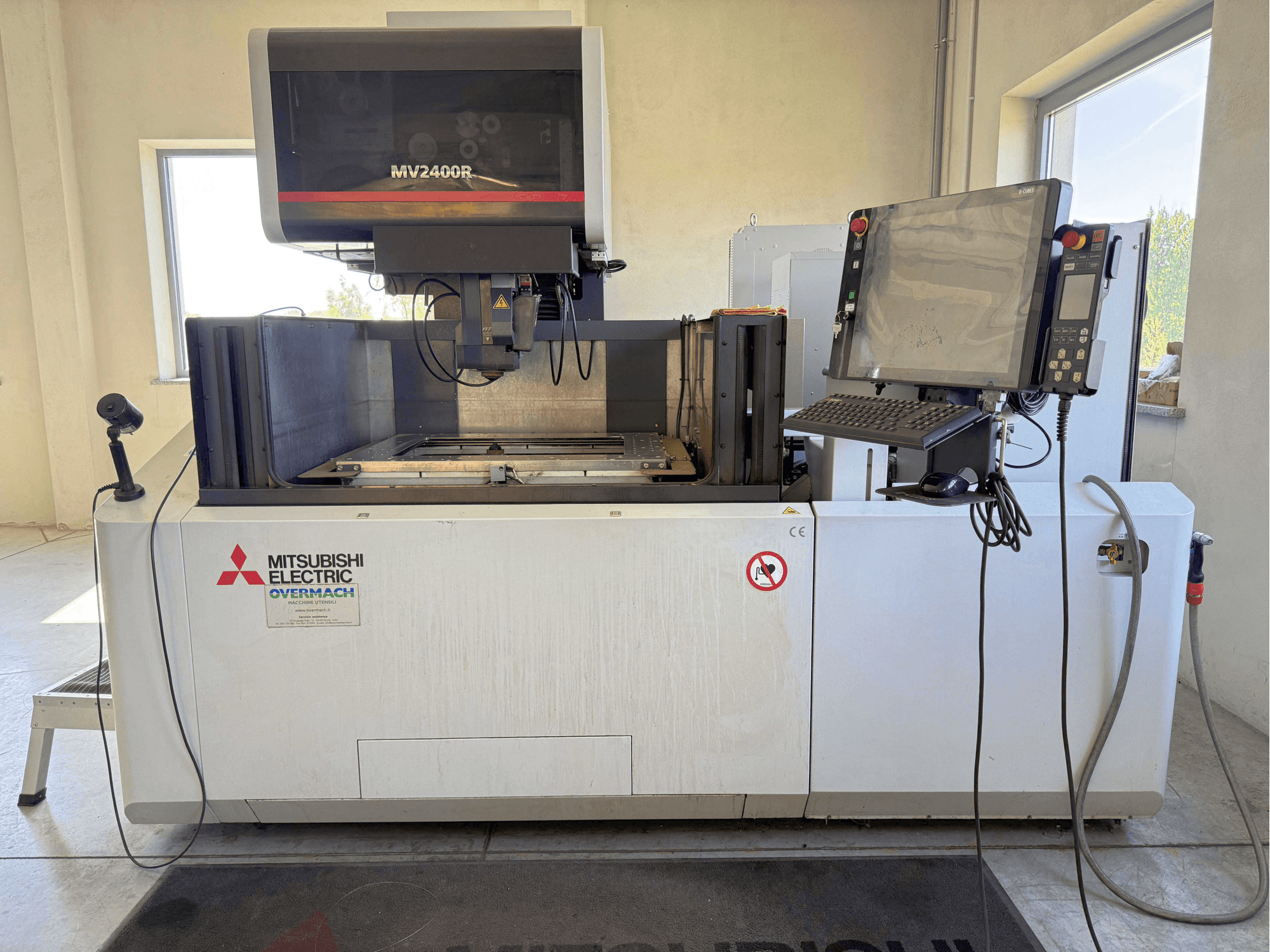 Mitsubishi MV2400R stiepļu elektroerozijas mašīna, skats no priekšpuses, kurā redzams vadības panelis, darba zona un drošības zīmes.