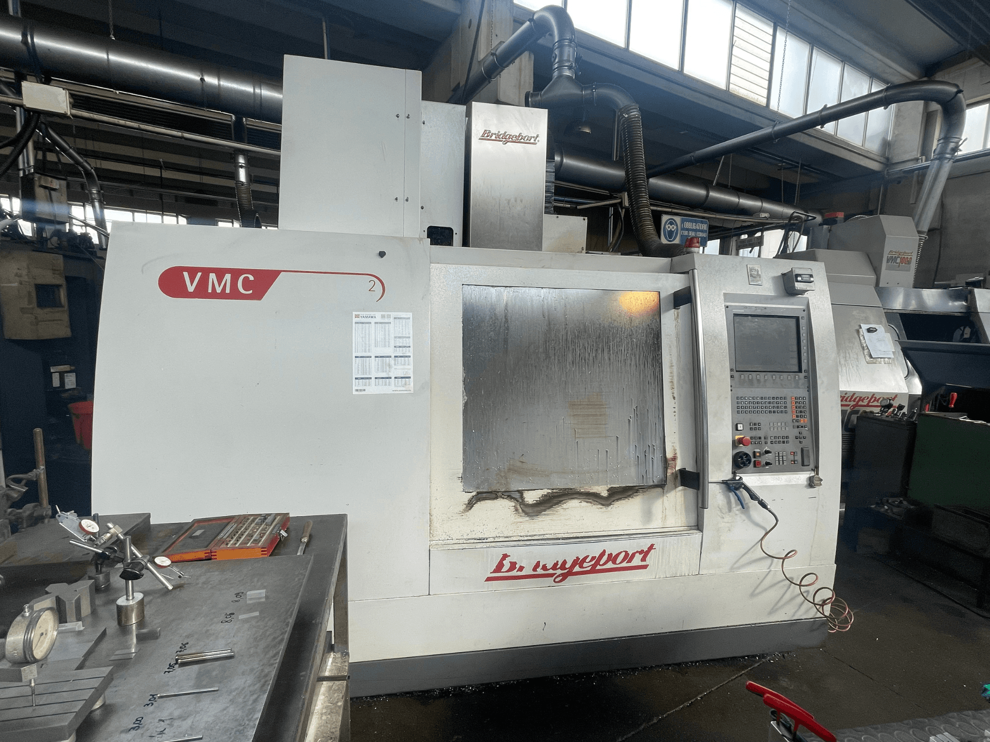 Vertikālais apstrādes centrs „Bridgeport VCM 800-30” darbnīcā, kurā redzams vadības panelis un instrumentu galds. Skats no priekšpuses ar redzamām detaļām.