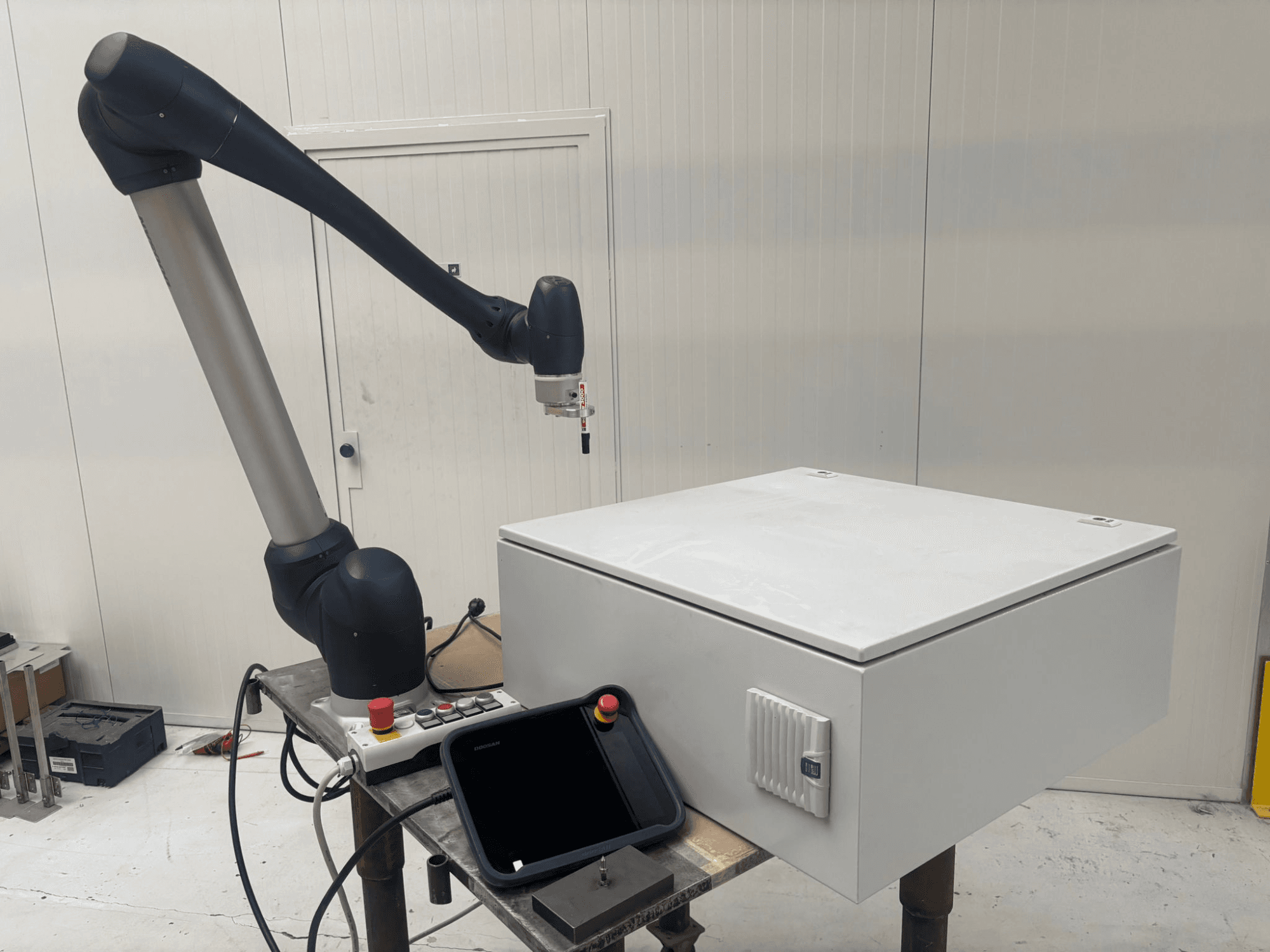 DOOSAN M0617 kolaboratīvais robots sānskatā, ar izstieptu robota roku virs vadības saskarnes un baltas kastes uz galda.
