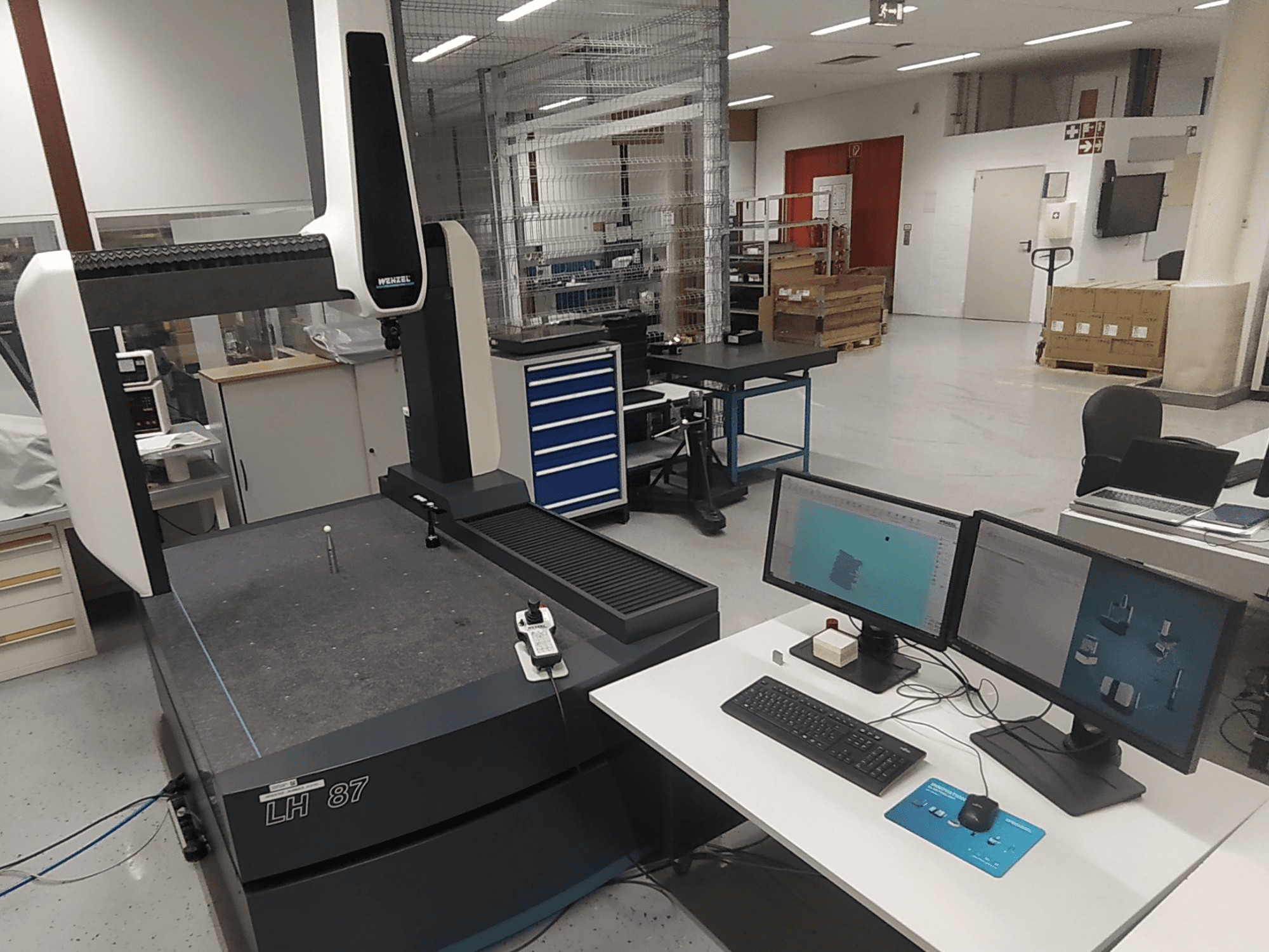 Wenzel LH 87 koordinātu mērīšanas iekārta, kas redzama laboratorijā, ar diviem monitoriem un mērīšanas zondi uz galda.