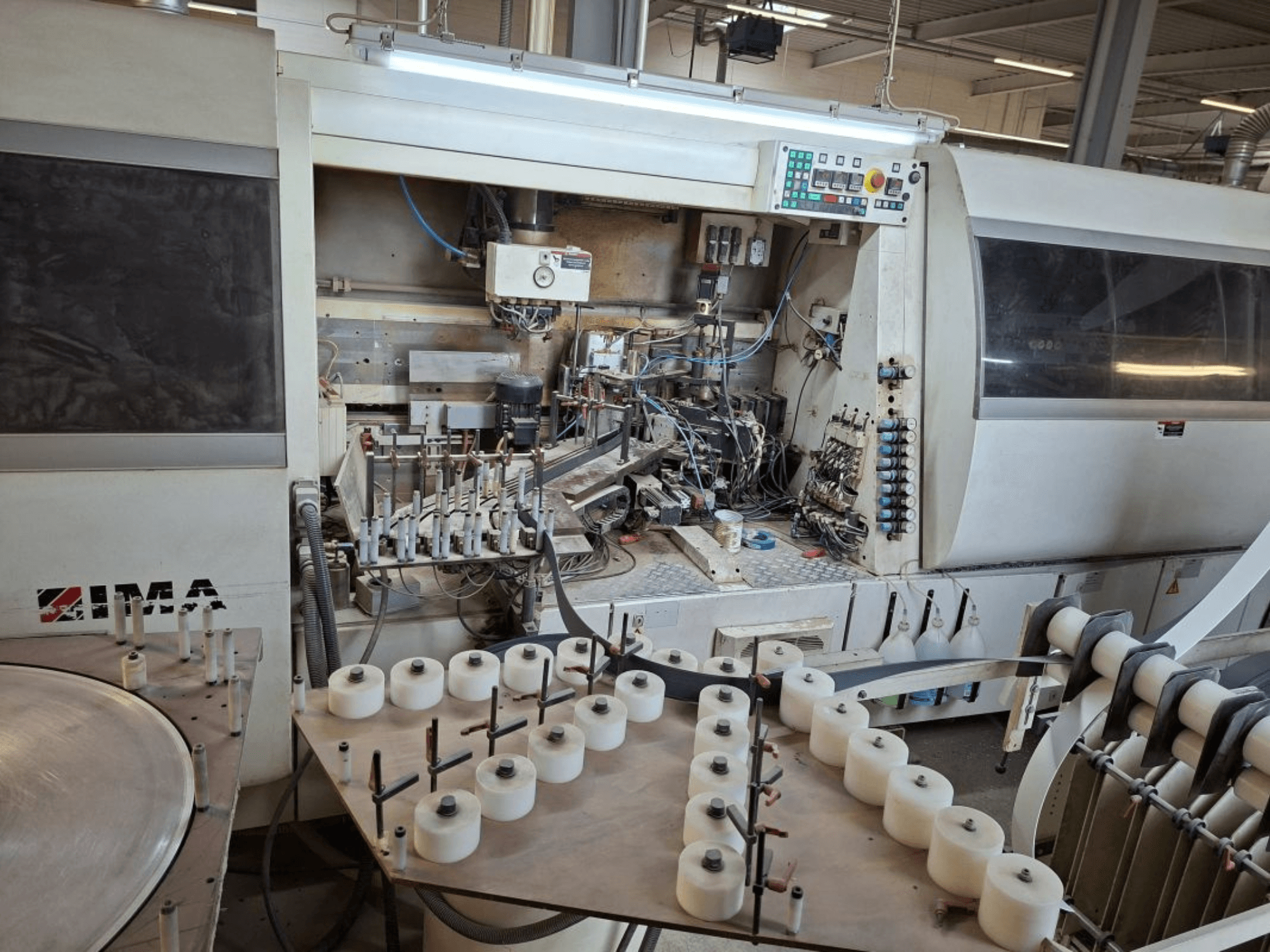 IMA iekārtas, skatoties no sāniem, ar sarežģītām mehāniskām detaļām un apaļu galdu ar baltām spoles.