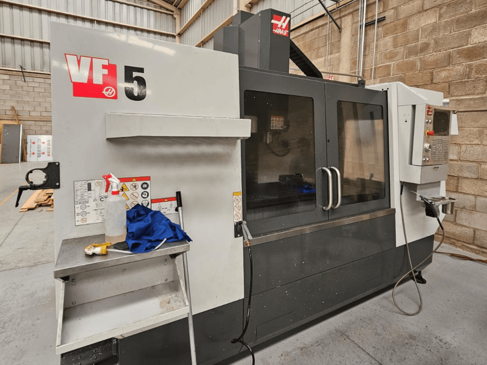 Haas VF-5 CNC frēzmašīna priekšpusē, ar vadības paneli, dzesēšanas šķidruma pudeli, zilu drānu un drošības instrukcijām.