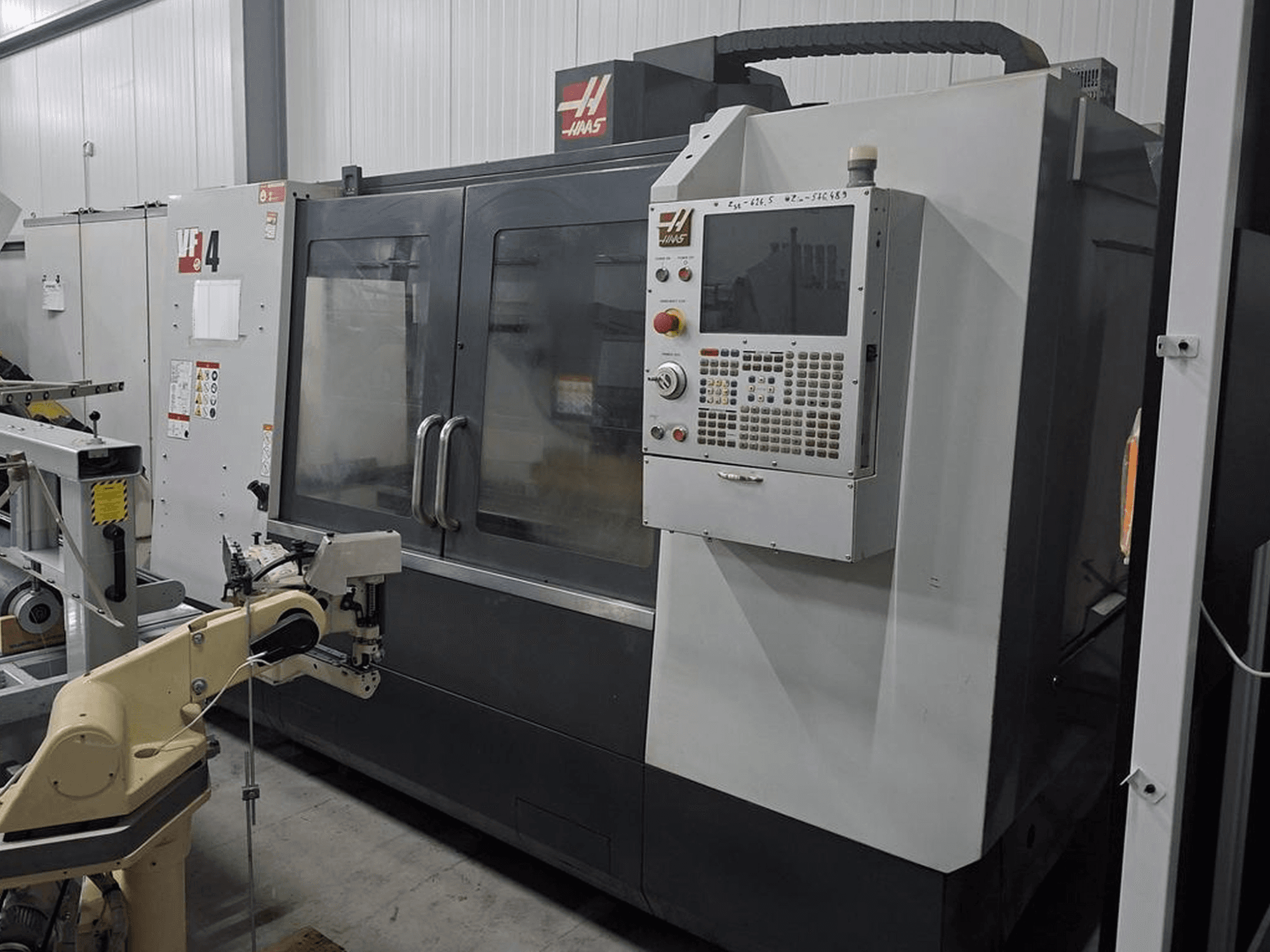 Haas VF-4 CNC frēzmašīna priekšpusē ar ciparu vadības paneli un robotizētu roku automatizācijai.
