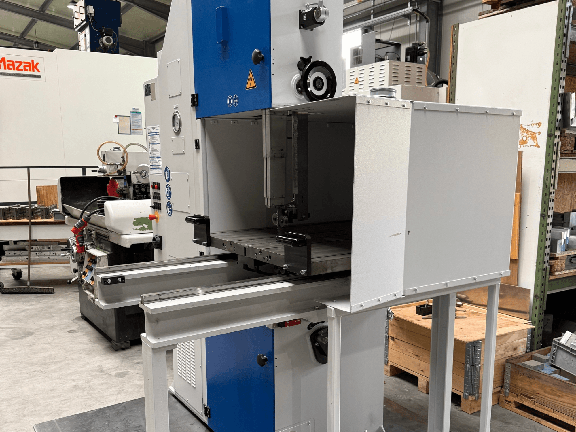 Mazak industriālā mašīna, skatoties no priekšpuses, ar zilu vadības paneli, metāla galdu un drošības brīdinājumiem.