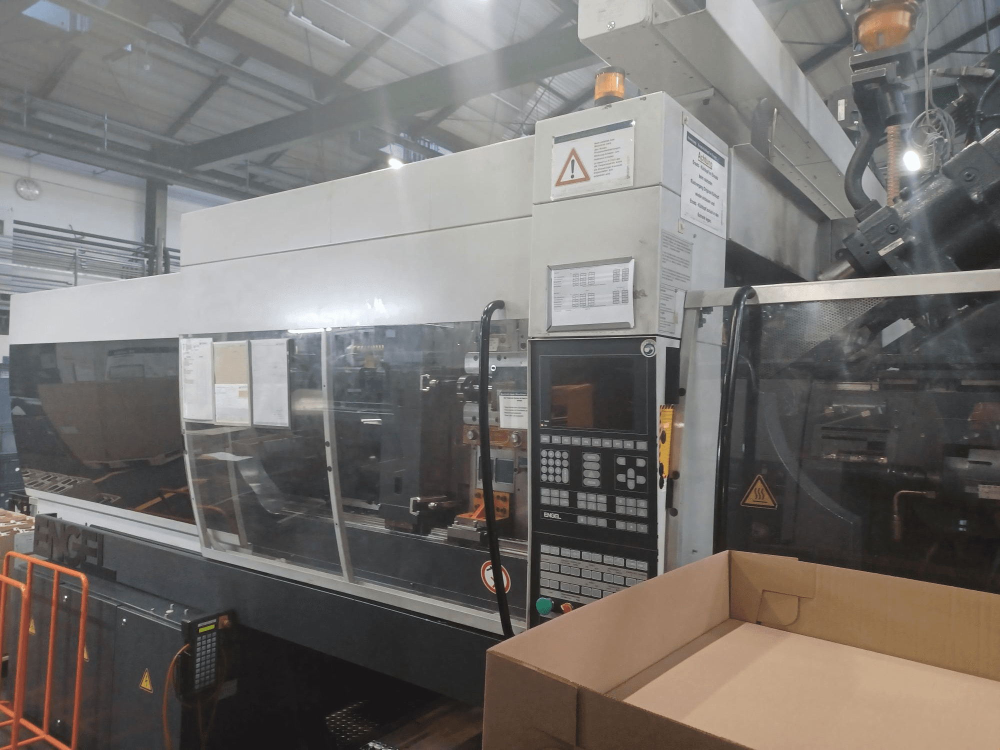 Engel iesmidzināšanas veidošanas iekārta, skats no priekšpuses, ar vadības paneli, drošības etiķetēm un blakus novietotu tukšu kasti.