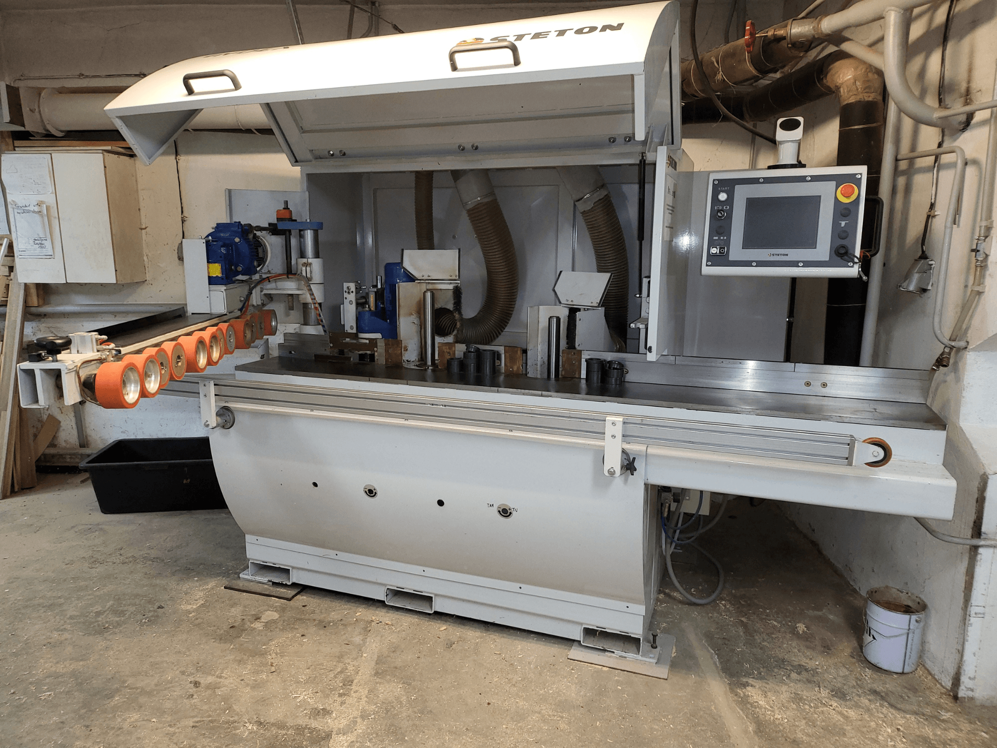 Stetton kokapstrādes mašīnas priekšējais skats ar vadības paneli, padeves caurulēm un dažādām koka detaļām uz galda.