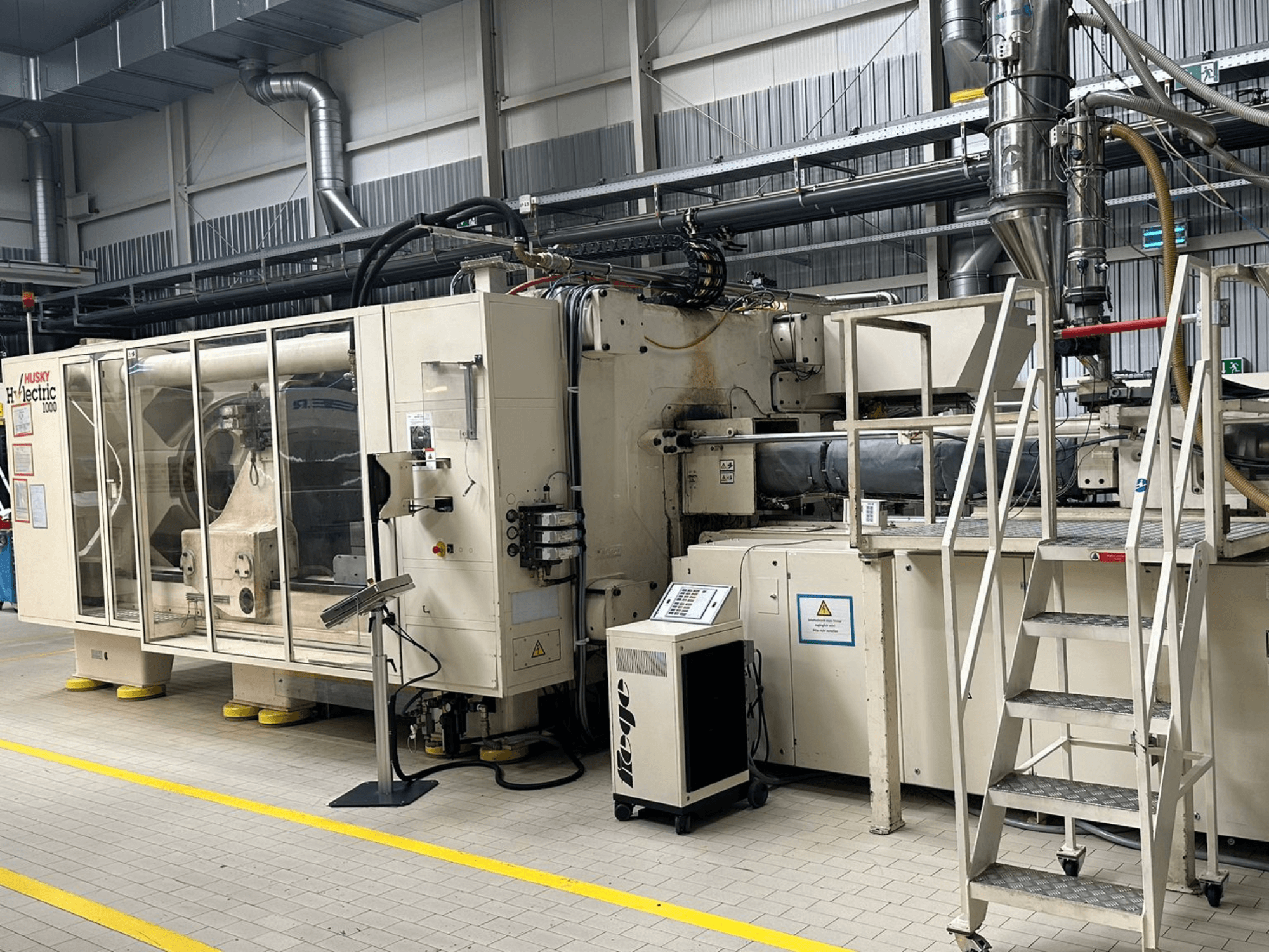 Husky Hylelectric H1000 RS95/80 elektriskās injekcijas formēšanas mašīnas priekšskats rūpnīcas vidē, kurā redzams vadības panelis un pakāpieni.