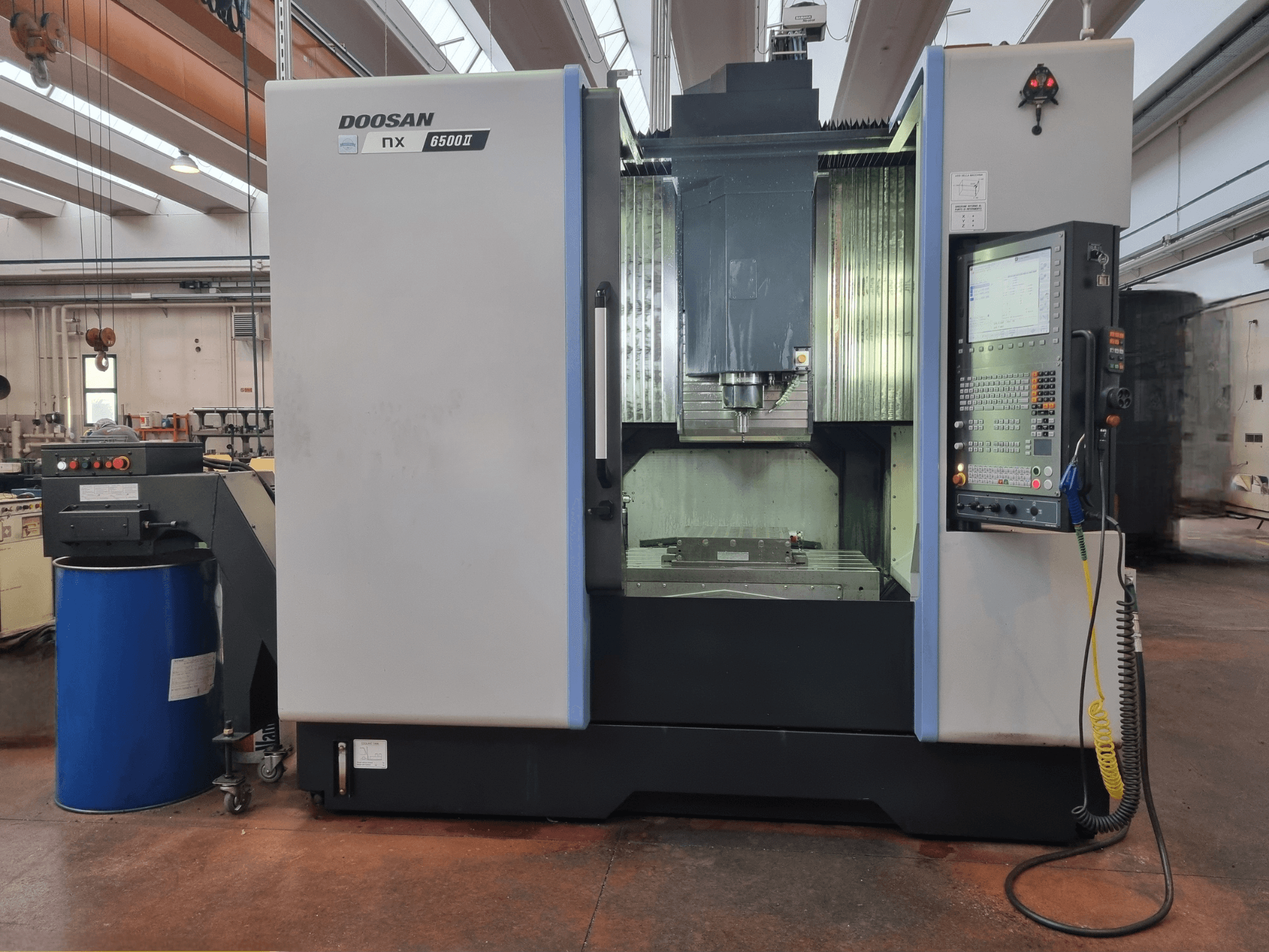 Doosan NX 6500 II CNC mašīna, skats no priekšpuses, ar vadības paneli un apstrādes zonu, kas novietota blakus zilai mucai.