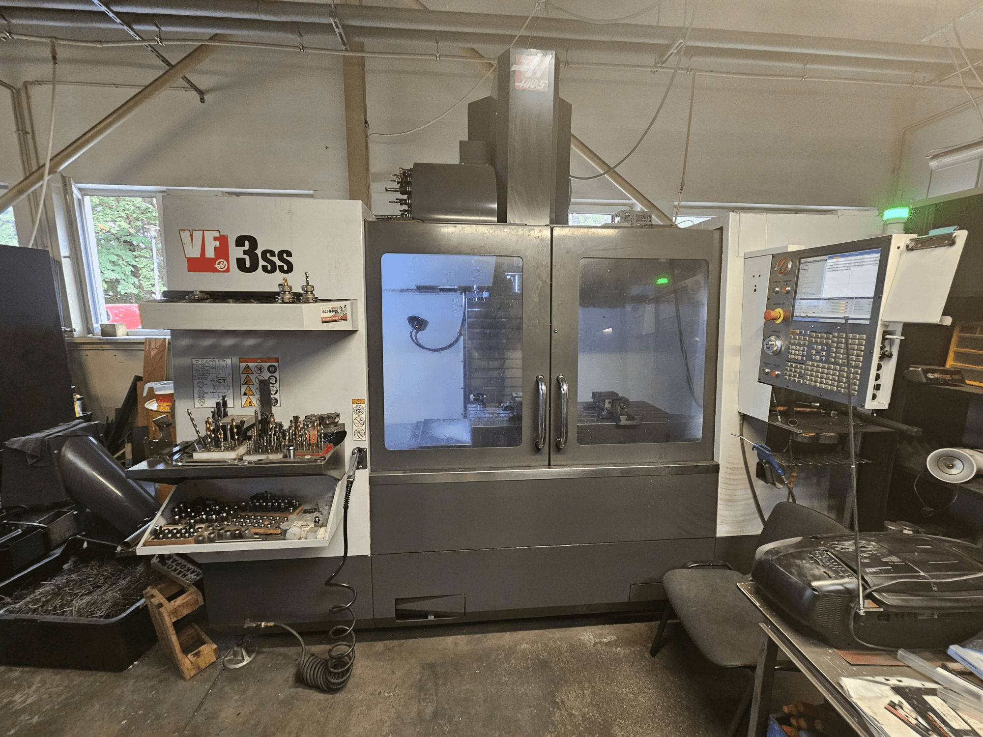 Vertikālais apstrādes centrs „Haas VF-3SS“ darbnīcā, skats no priekšpuses, kurā redzams vadības panelis un instrumentu glabāšanas nodalījums.