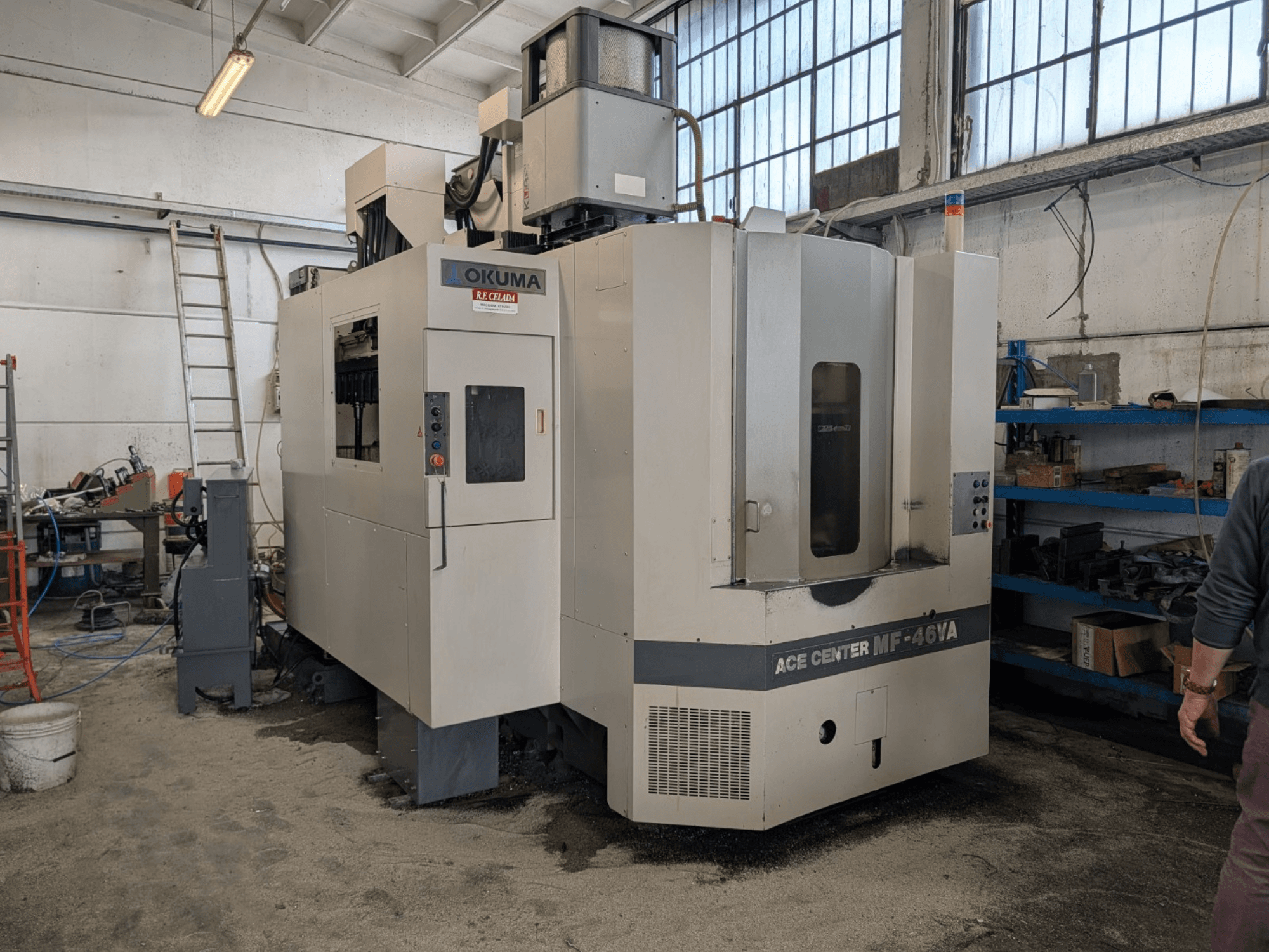 Okuma ACE CENTER MF-46VA vertikālais apstrādes centrs darbnīcā, priekšējais skats ar vadības paneli un apkārtējām iekārtām.