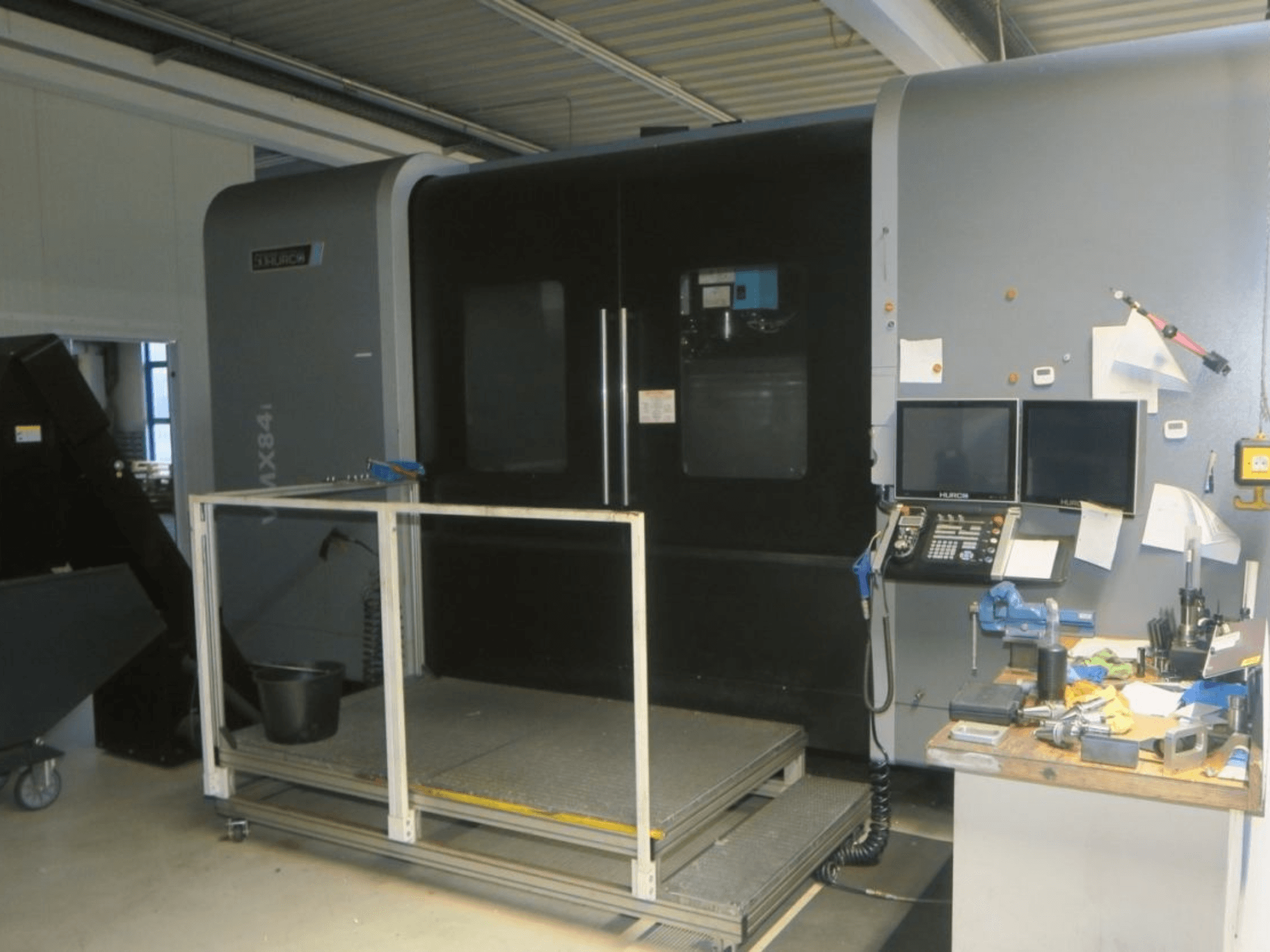 Hurco MX84 CNC darbgalds priekšpusē, kurā redzami divi monitori, vadības panelis un instrumentu darba vieta ar dažādiem instrumentiem.