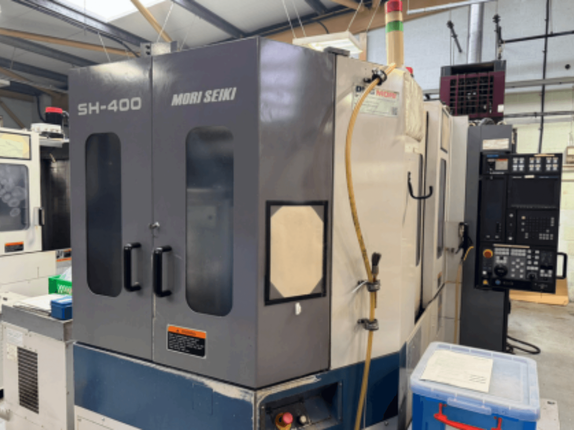 Mori Seiki SH-400 CNC darbgalds, skats no priekšpuses, ar divām durvīm, vadības paneli un piestiprināšanas šļūteni. Blakus atrodas zils glabāšanas konteiners.