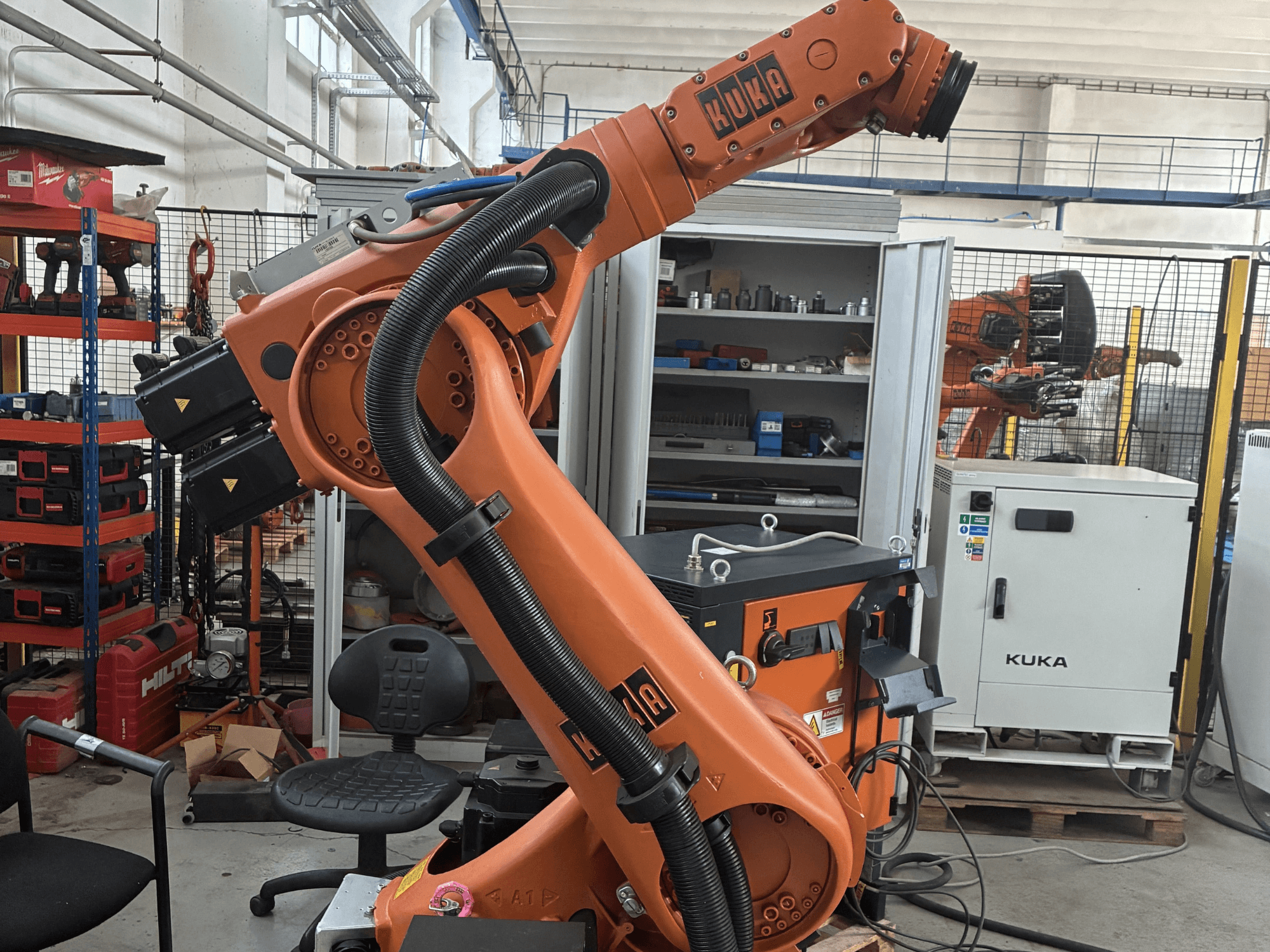 Robota roka KUKA KR30-3 rūpnieciskā vidē, skats no sāniem, kurā redzami kabeļi un vadības ierīces, fonā - instrumenti un plaukti.