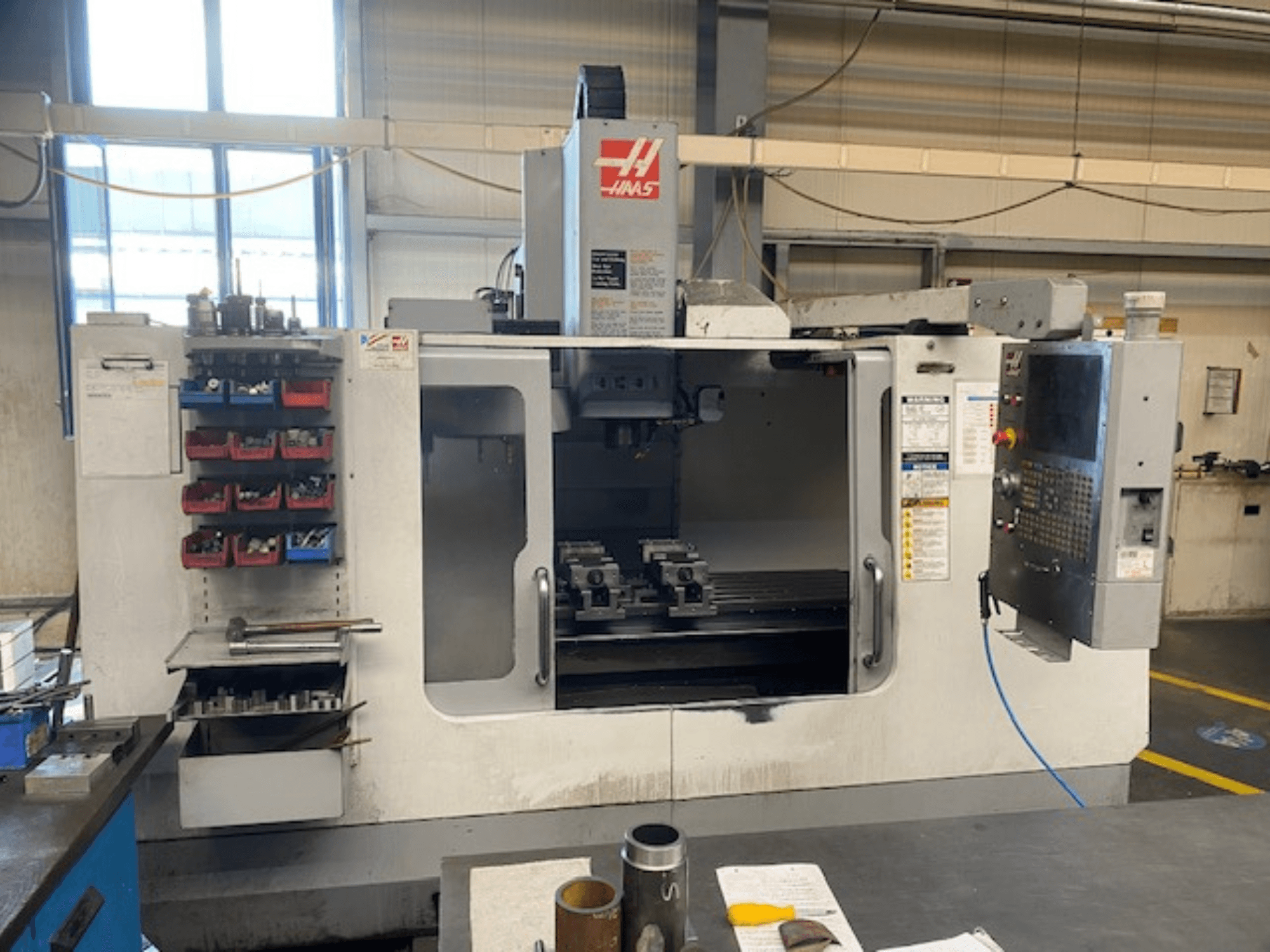 Haas CNC frēzmašīnas priekšējais skats, modelis VF-2, ar redzamu instrumentu glabāšanas vietu, vadības paneli un darba vietas instrumentiem.