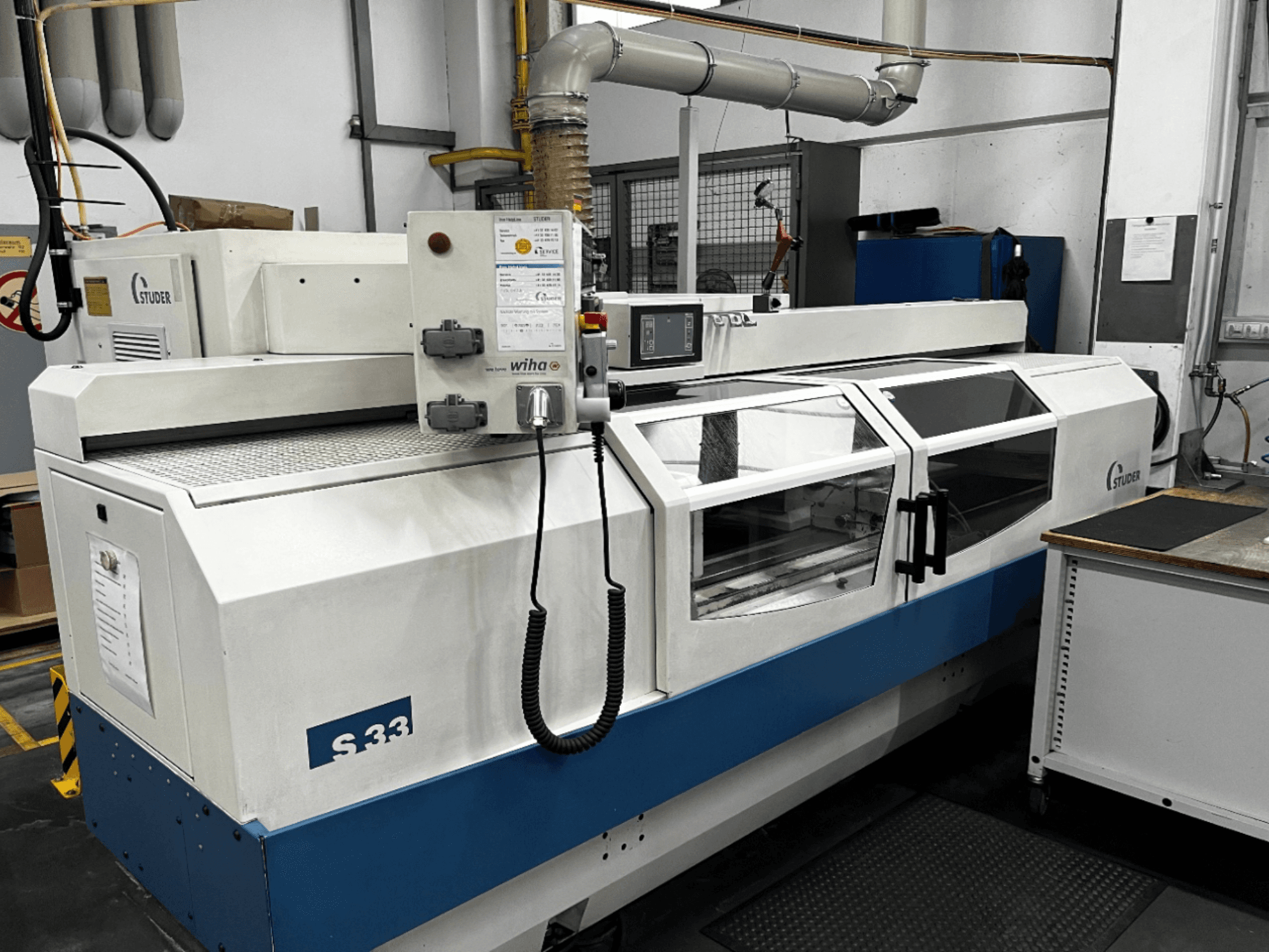 STUDER S33 CNC cilindriskās slīpmašīnas, skats no priekšpuses; vadības panelis un stikla drošības durvis. Rūpnieciskā vide.