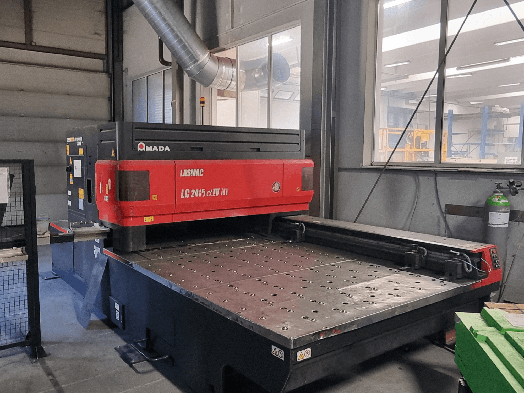 Amada LASMAC LC 2415 αⅡ NT lāzera griešanas iekārta sarkanā un melnā krāsā, skats no priekšpuses, ar motorizētu griešanas gultni un precīzām vadības ierīcēm.
