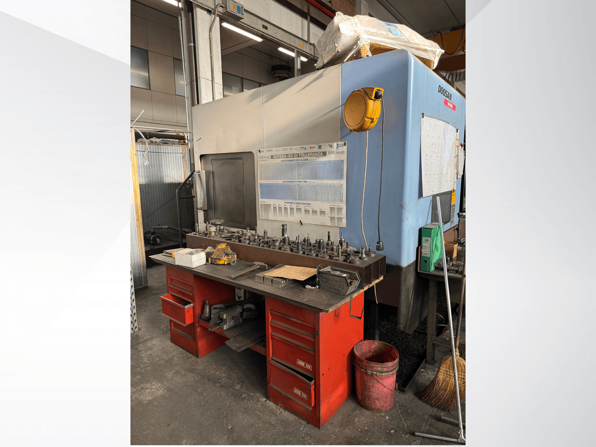 Doosan apstrādes centrs ar sarkanu darbagaldu un noliktavu, kurā izvietoti instrumenti un aprīkojums, skatīts no priekšpuses.