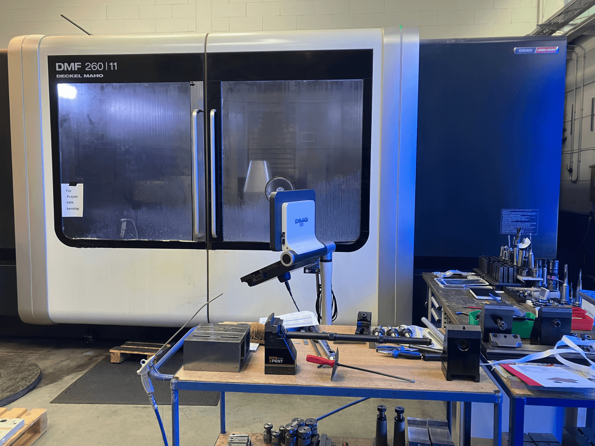 DMG DECKEL MAHO DMF 260|11 vertikālā apstrādes centra DMG DECKEL MAHO DMF 260|11 priekšējais skats ar instrumentiem un materiāliem uz darbstacijas.