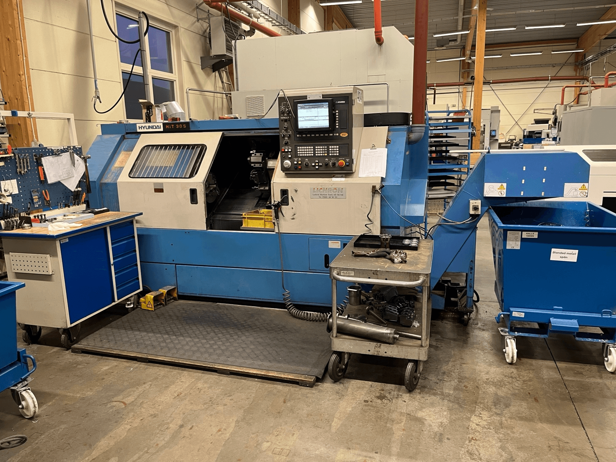 Hyundai HIT 30S horizontālās virpošanas iekārta, skats no priekšpuses, ar vadības paneli un instrumentiem uz blakus novietota ratiņa darbnīcā.