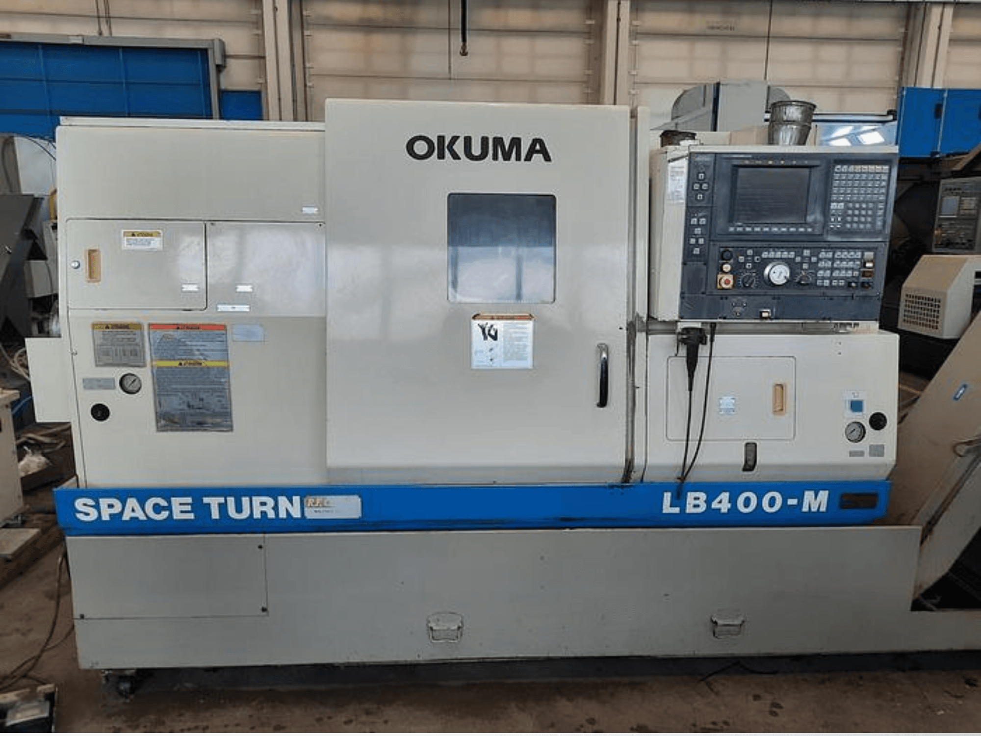 Okuma LB400-M CNC virpmašīna, priekšējais skats ar vadības paneli, brīdinājuma uzlīmes un redzams zils "SPACE TURN" zīmols.