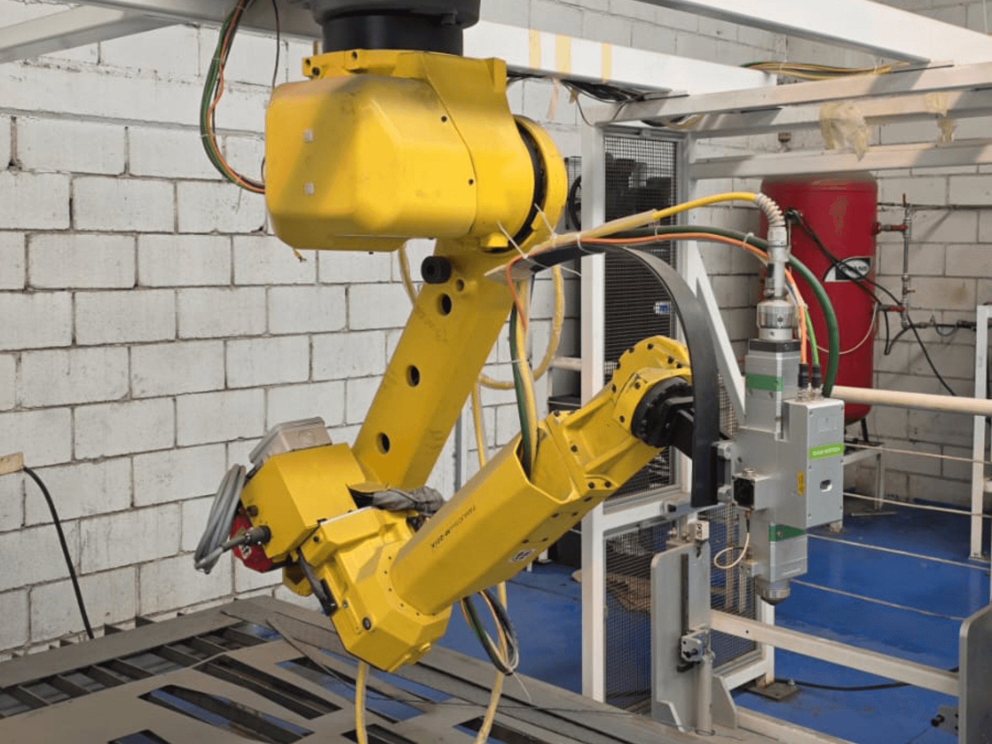 Dzeltena robotizēta roka ar precīzu instrumentu, kam ir vairāki savienojumi un kabeļi, skatīta no priekšējā leņķa rūpnieciskā vidē.