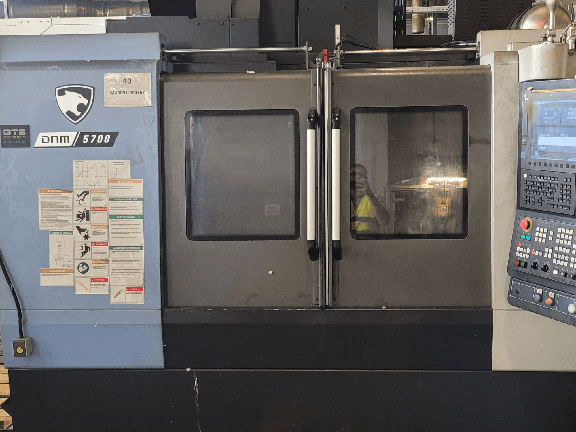 DOOSAN DNM 5700 vertikālā apstrādes centra skats no priekšpuses ar vadības paneli, drošības uzlīmēm un nedaudz pavērtām durvīm.