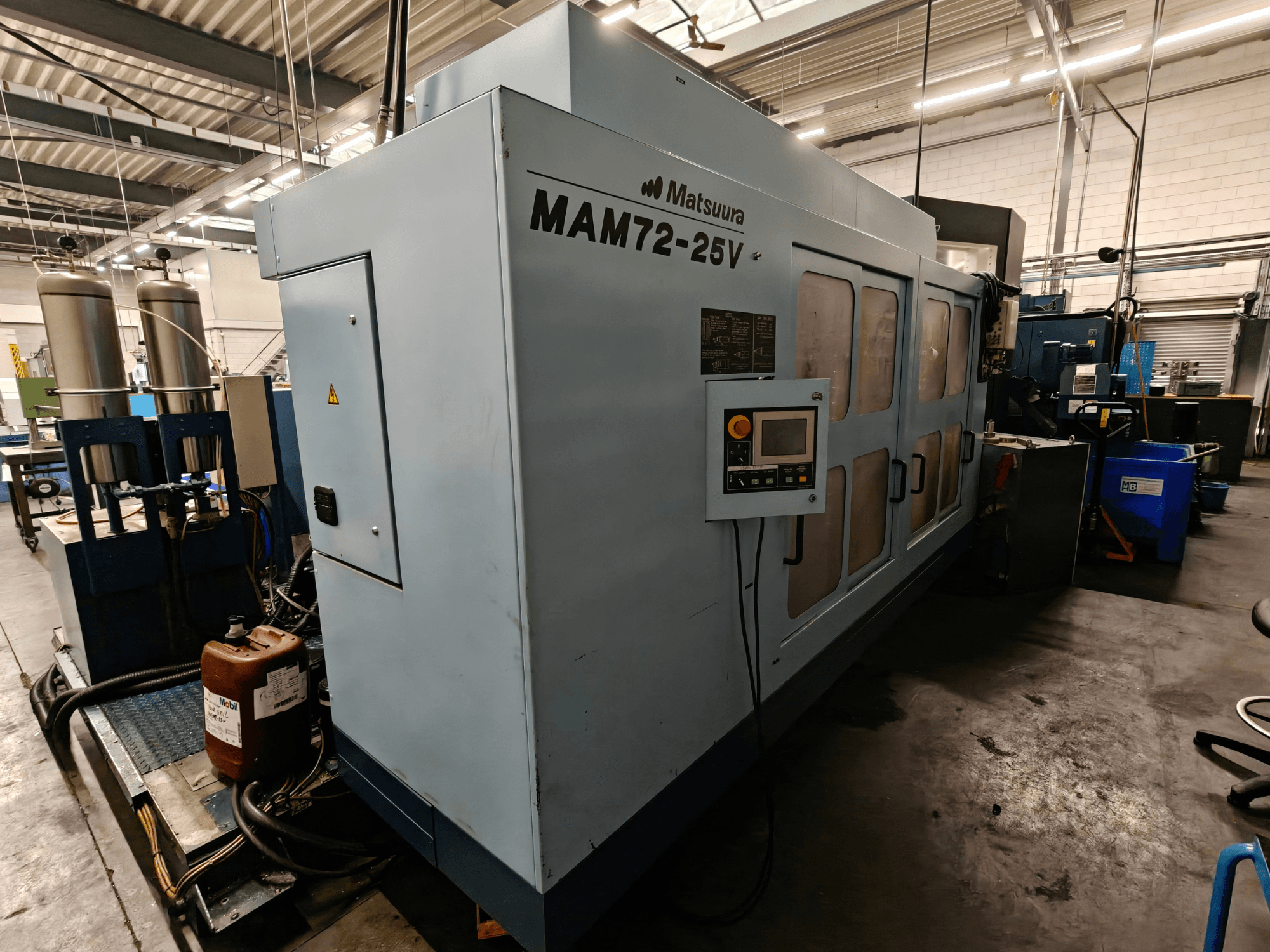 Matsuura MAM72-25V CNC darbgalds, skats no sāniem, ar vadības paneli, divām dzesēšanas šķidruma tvertnēm un darba vietu industriālā vidē.