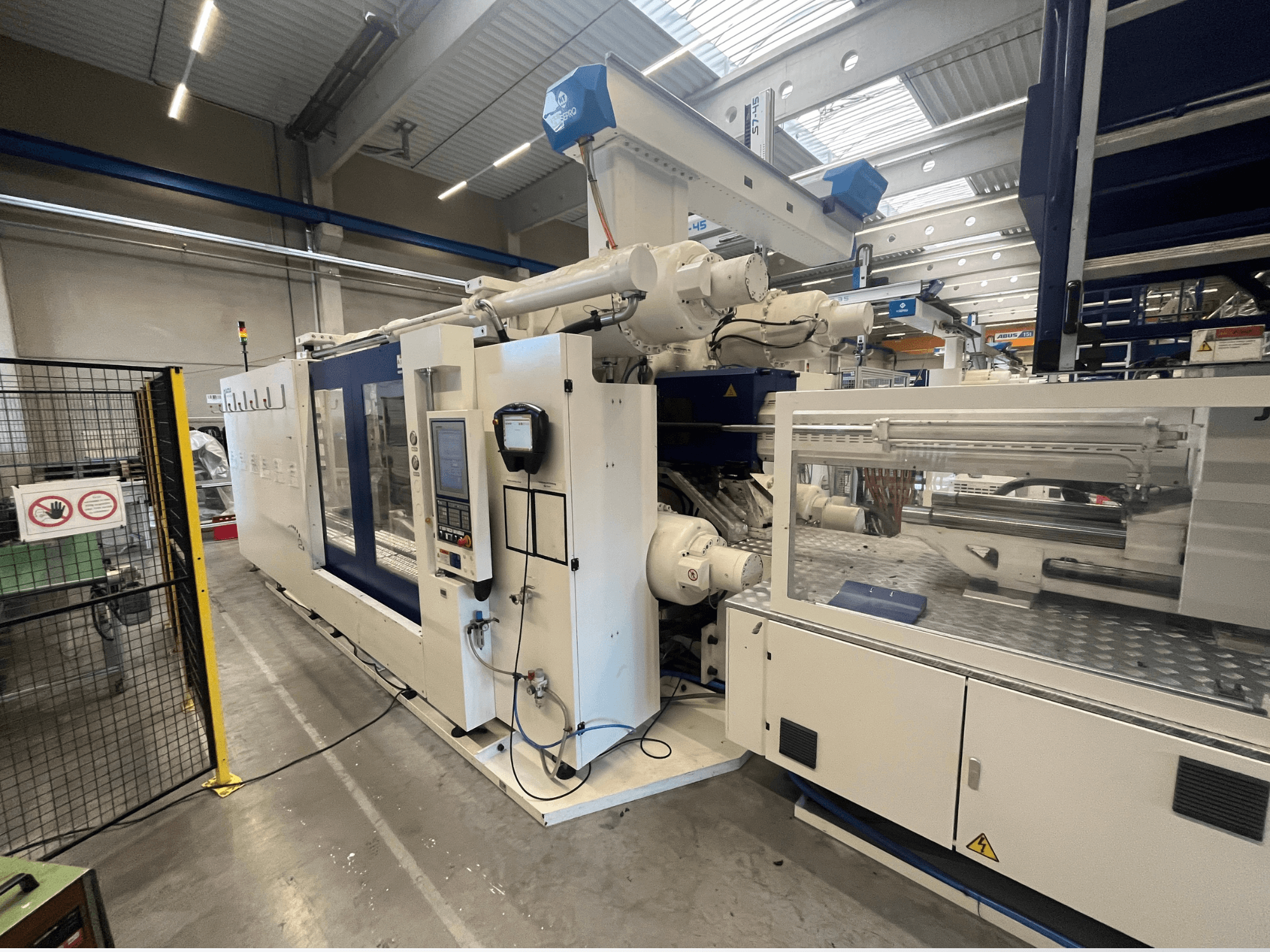 Baltas un zilas krāsas injekcijas formēšanas iekārta, skats no priekšpuses, ar vadības paneli, robotizēto roku un drošības zīmēm.