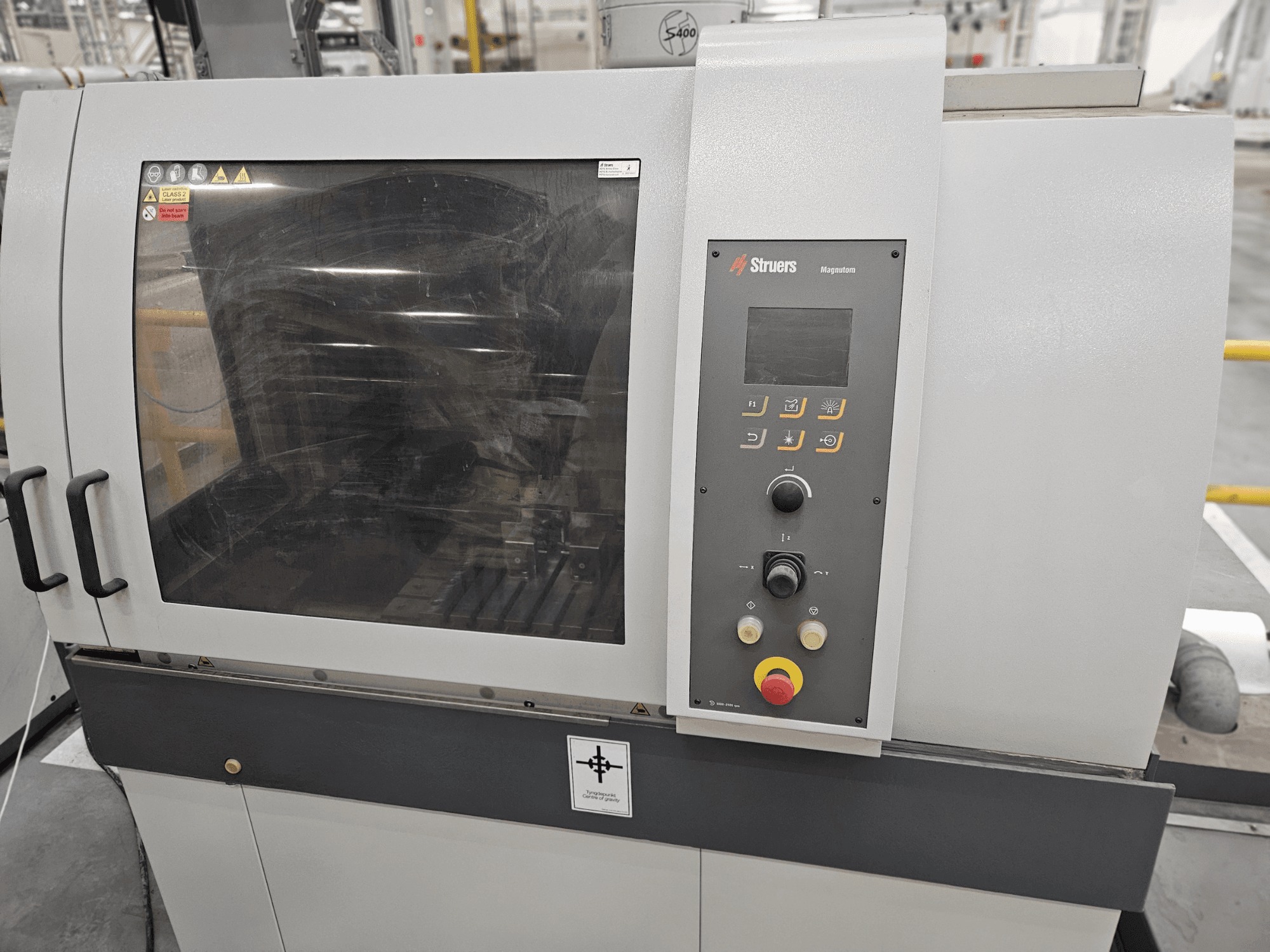 Struers Magpentor mašīnas priekšējais skats ar vadības paneli, pogām un lielu skatu logu. Redzamas tehniskās etiķetes.