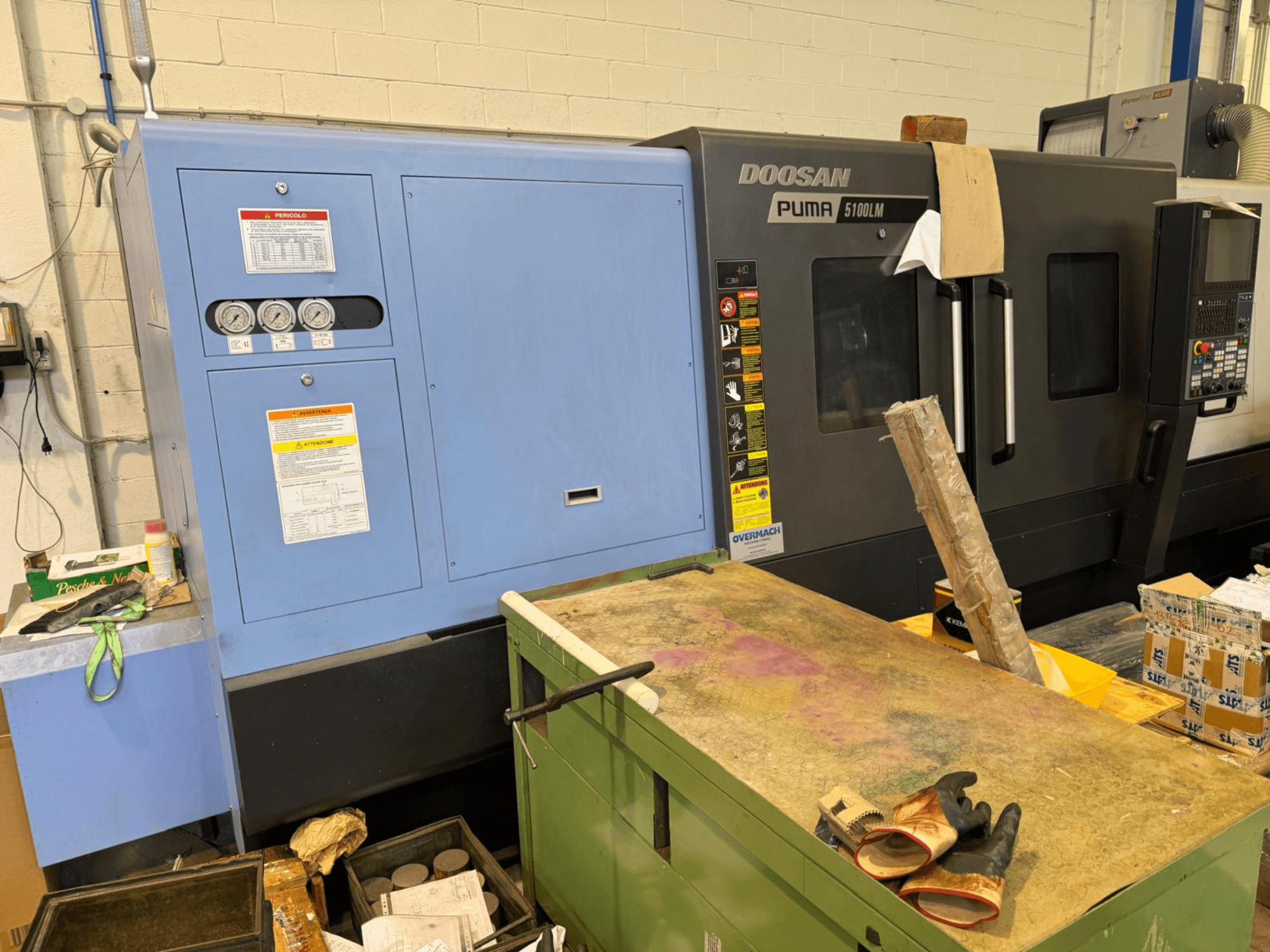 Doosan Puma 5100LM CNC mašīna un vadības panelis, skats no sāniem. Uz zilā vadības bloka redzami mērinstrumenti un brīdinājuma uzlīmes.