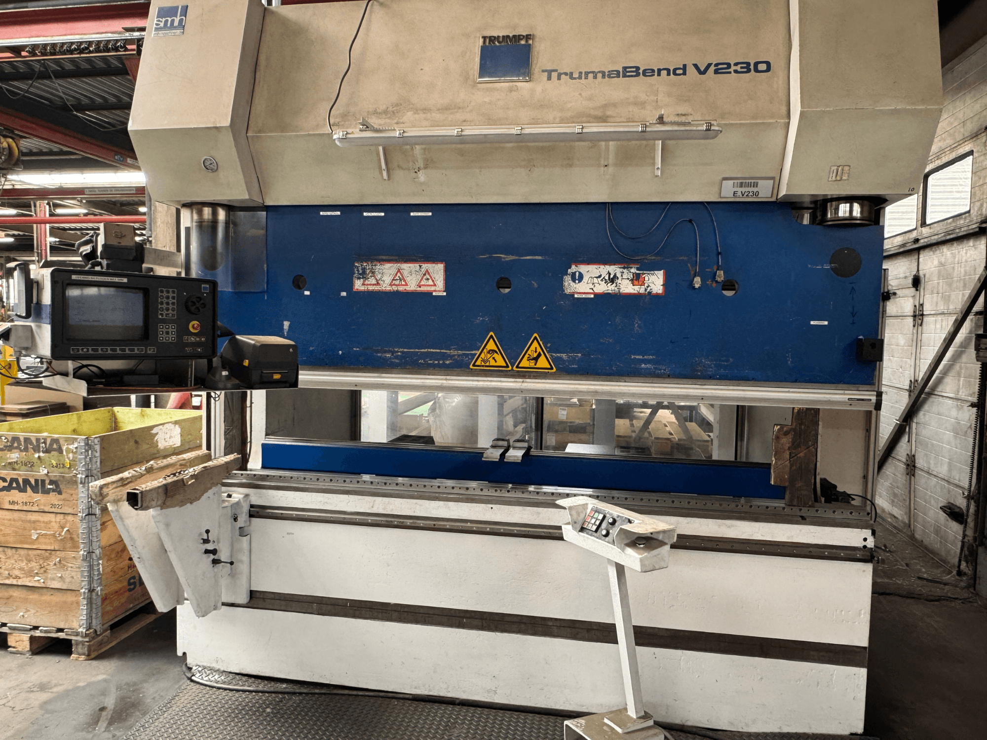 TRUMPF TrumaBend V230 preses bremze, skats no priekšpuses ar redzamu vadības paneli, brīdinājuma uzlīmes un apstrādājamās detaļas atbalsta roku.