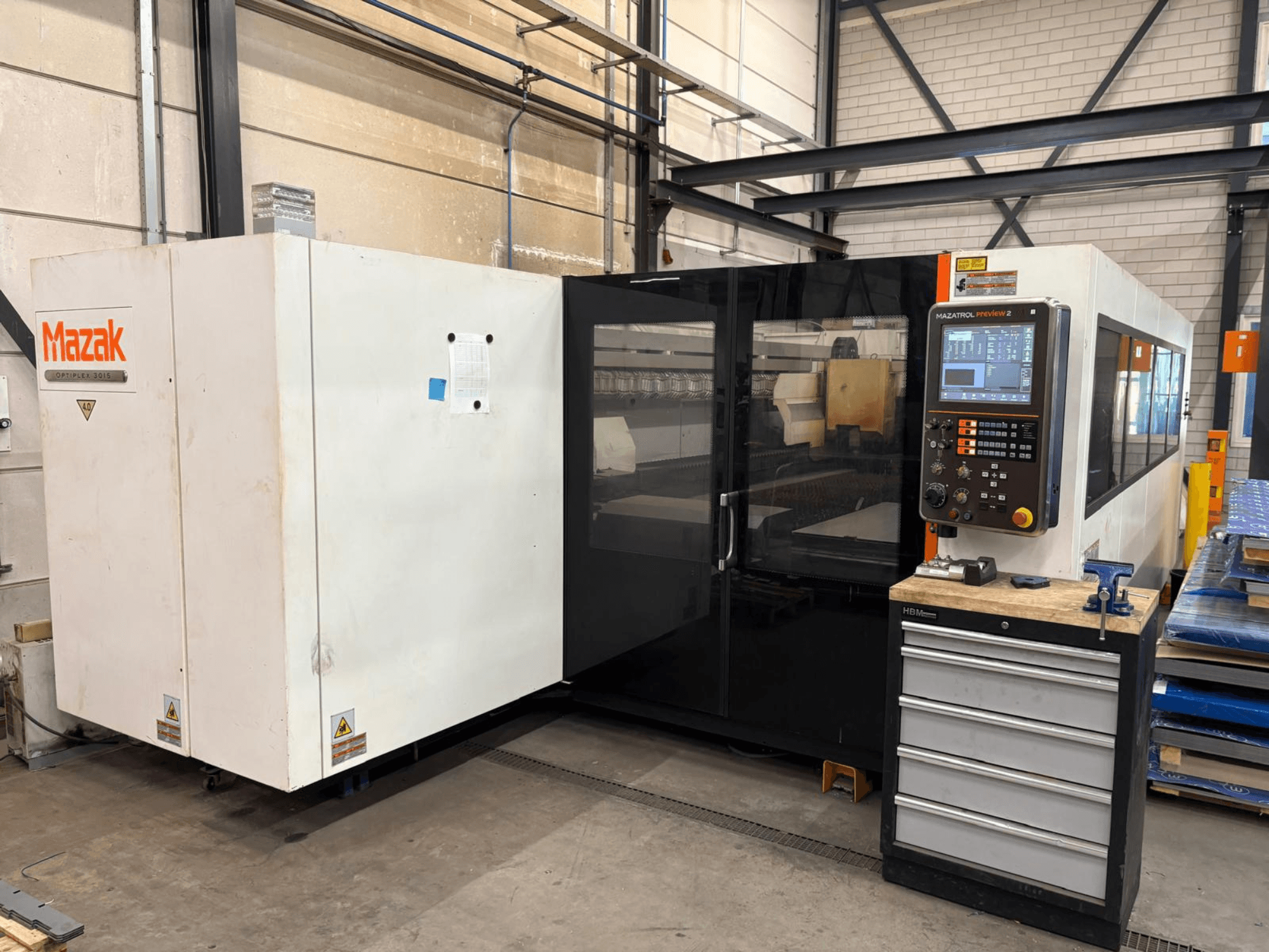 Mazak Optiplex 3015 CO₂ lāzergriešanas iekārta, skats no priekšpuses, kurā redzams vadības panelis un rūpnieciskā uzstādīšana.