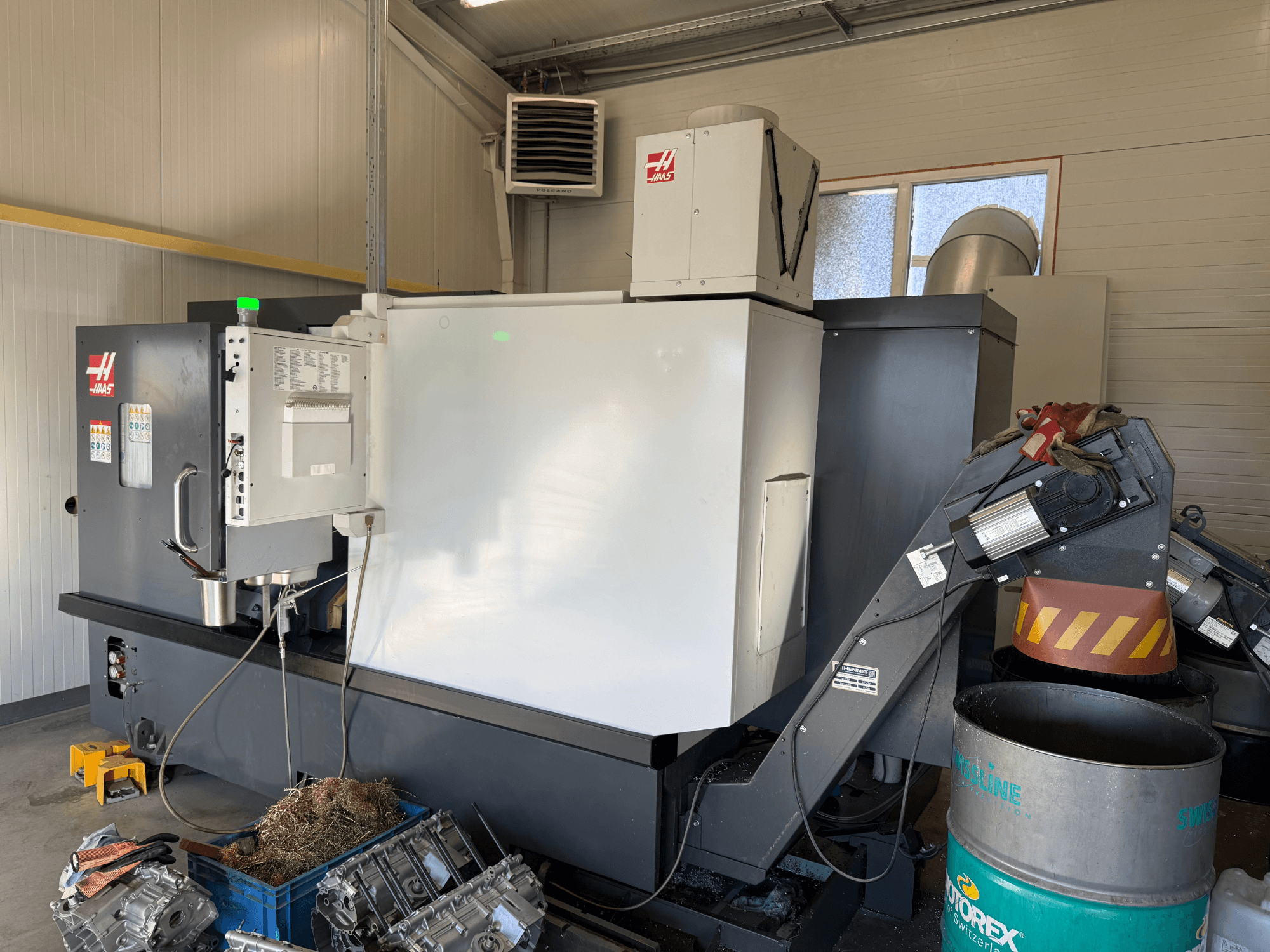 Haas CNC darbgalds ar vadības paneli, skats no sāniem; redzamas dzesēšanas šķidruma un eļļas caurules; metāla atgriezumi zilā tvertnē; rūpnieciska vide.