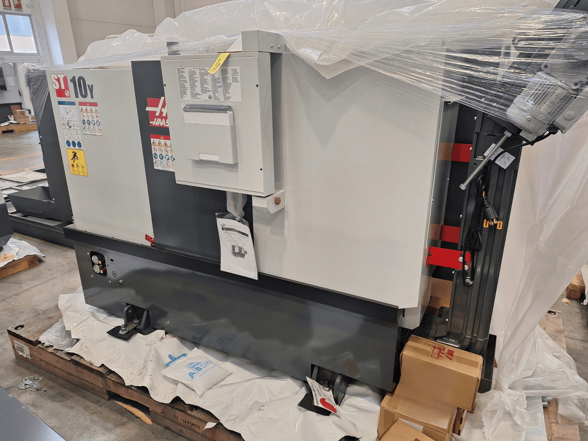HAAS ST-10Y EU horizontālās virpošanas mašīnas sānskats, ietināta plastmasā, redzams vadības panelis un brīdinājuma uzlīmes.