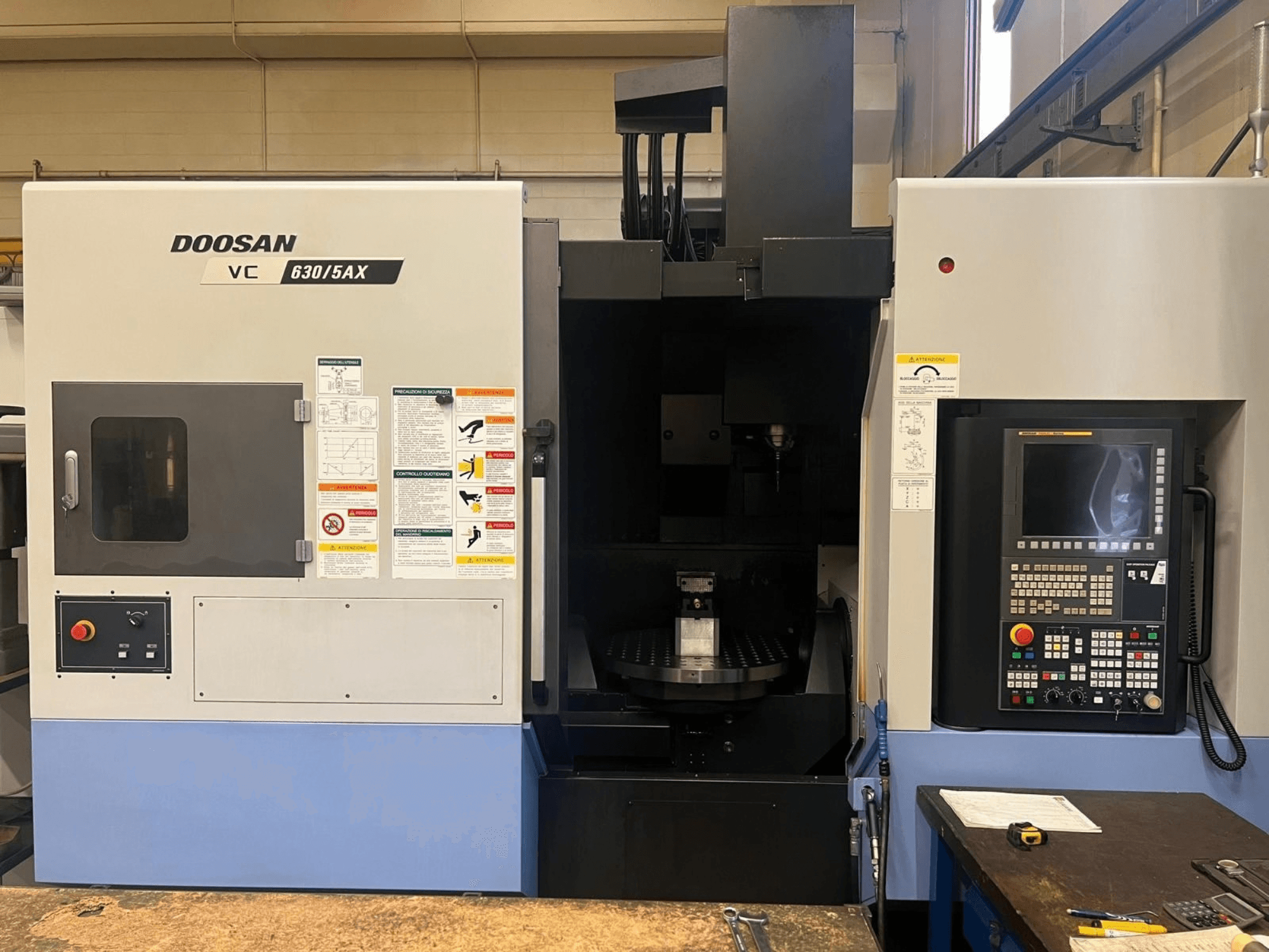 DOOSAN VC 630/5AX vertikālā apstrādes centra skats no priekšpuses, kurā redzams vadības panelis un vārpstas zona ar redzamām drošības uzlīmēm.