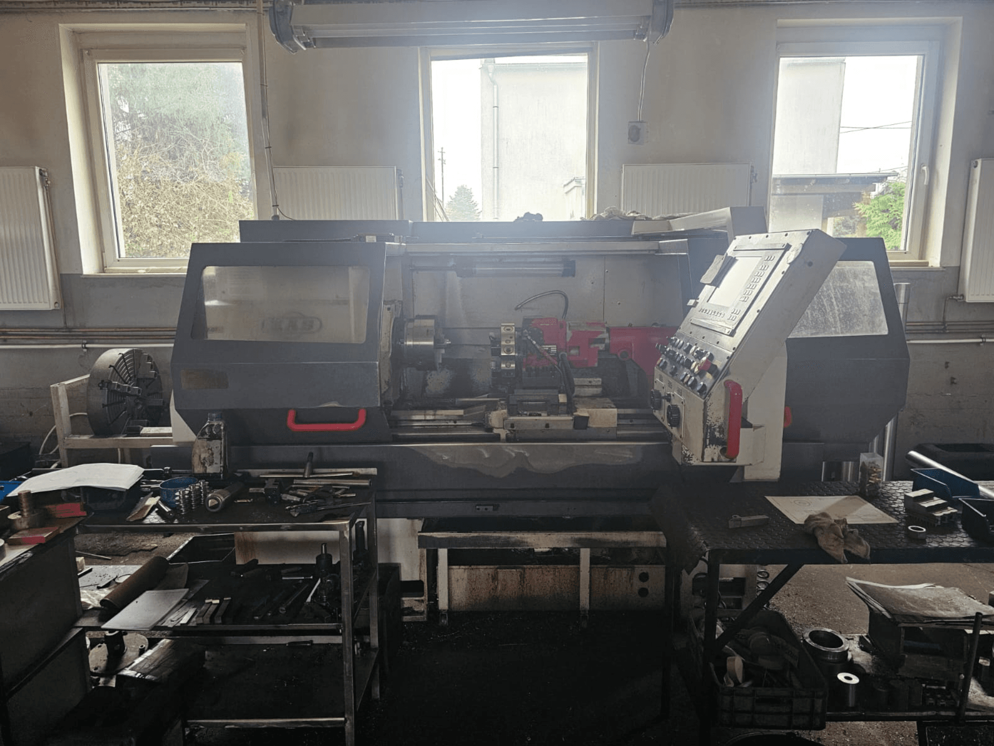 Virpas darbnīcā, skats no priekšpuses, ar vadības paneli un instrumentiem uz galdiem, logu ieskauj logi un pārblīvēta darba telpa.