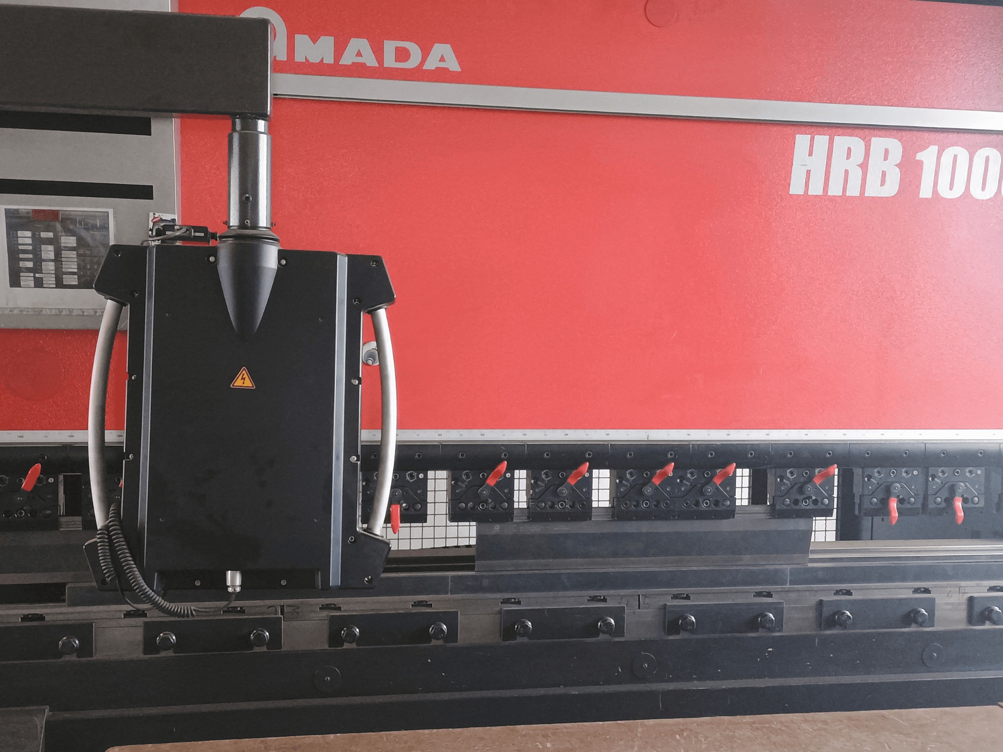 Amada HRB 100 liekšanas mašīnas priekšējais skats; ar melnu vadības galvu ar konusveida sprauslu un regulējamām skavām.
